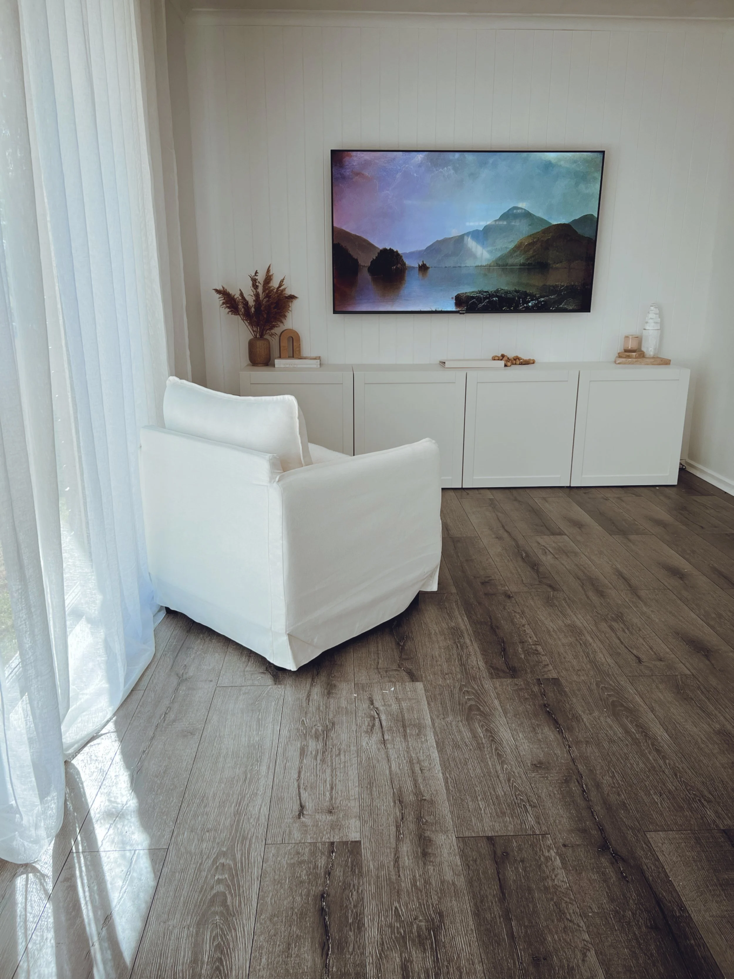 A living room with a white armchair, white cabinet, a wall-mounted TV displaying a nature scene with mountains and water, and decorative items on the cabinet, with sunlight coming through sheer curtains.