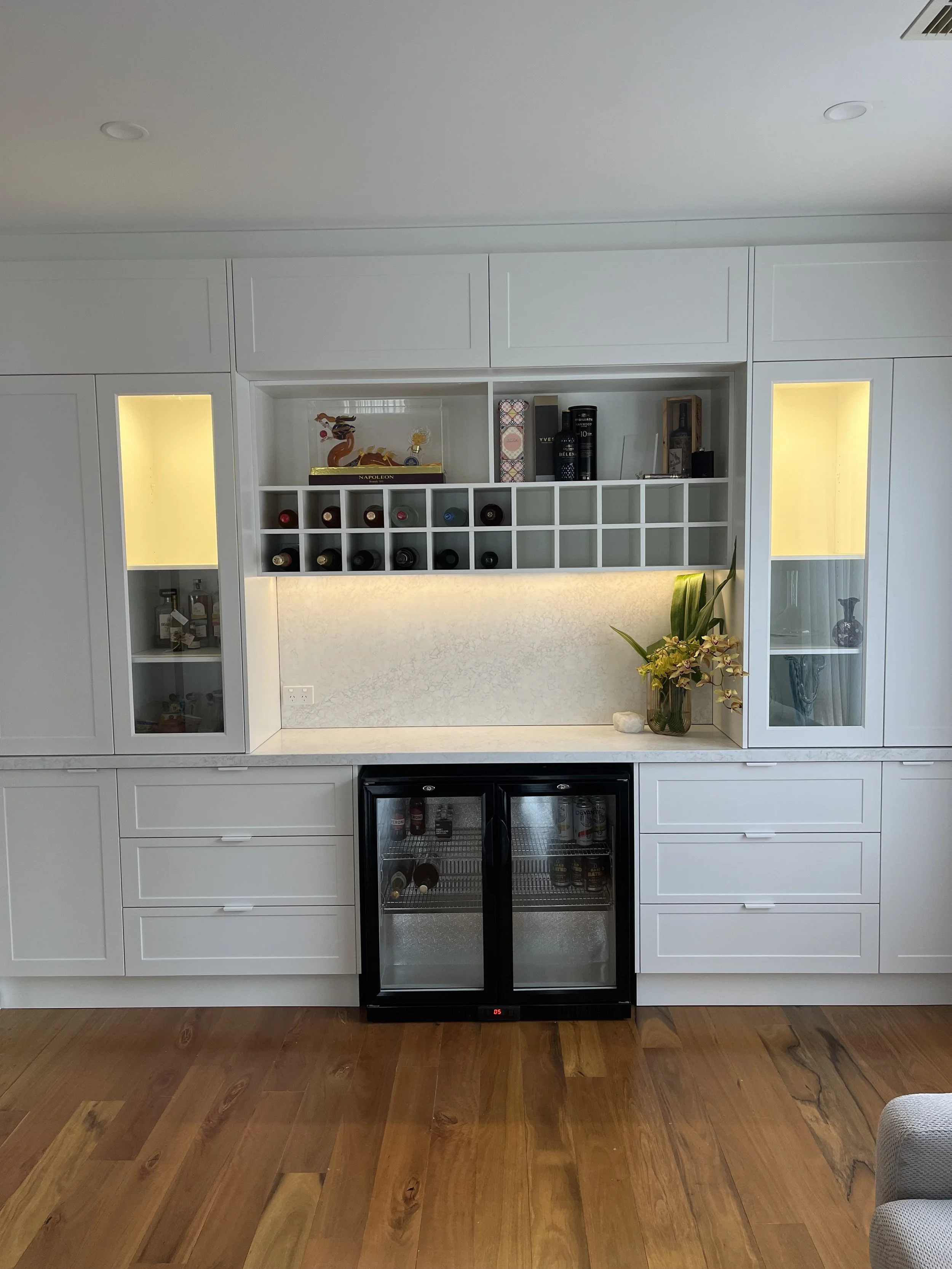 A modern built-in bar area with white cabinets, wine bottles in a rack, glass-front refrigerator, and decorative items including plants and books, on a wooden floor.