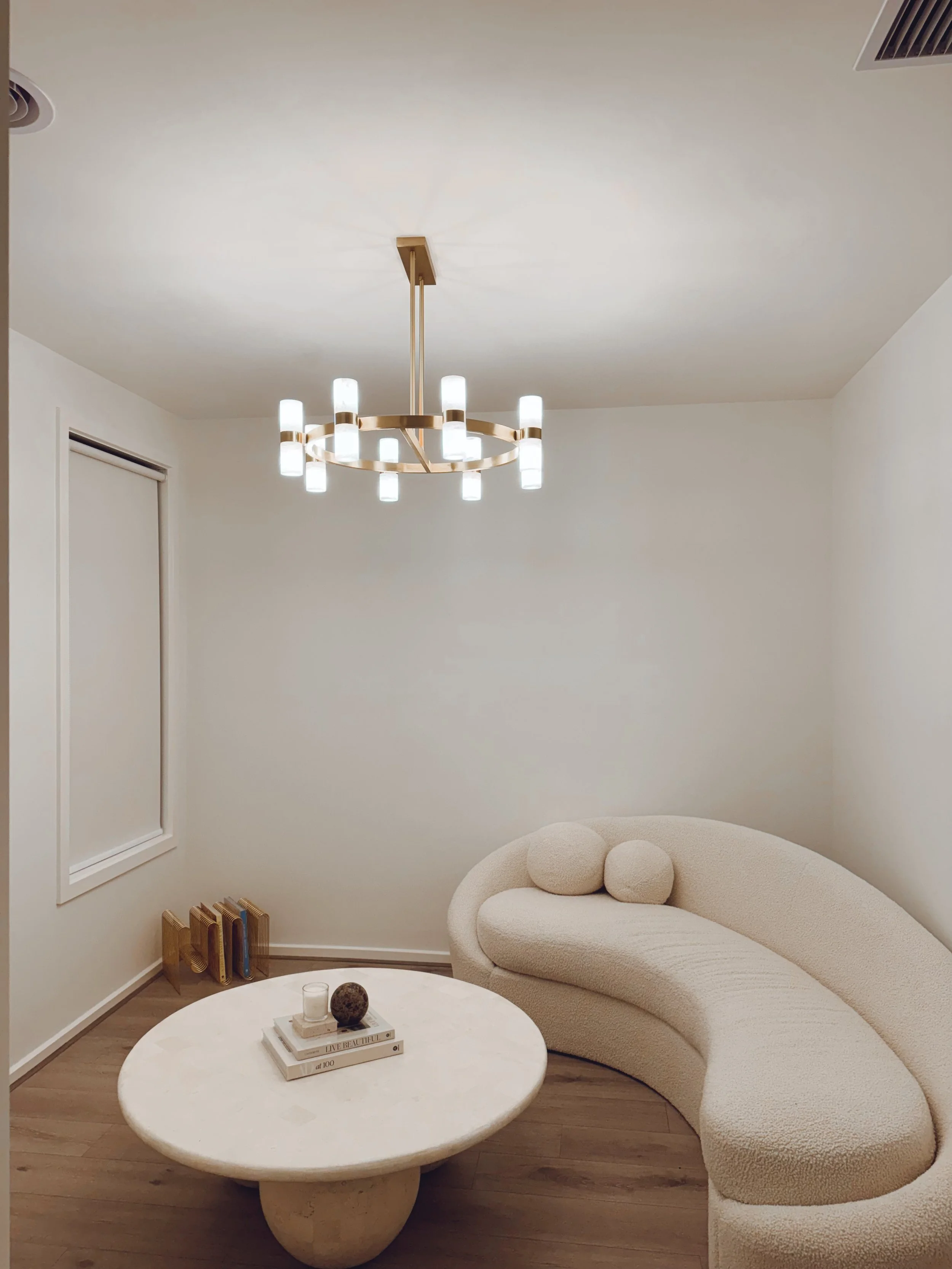Minimalist living room with a cream curved sofa, round marble coffee table, modern chandelier, and neutral decor on a wooden floor.