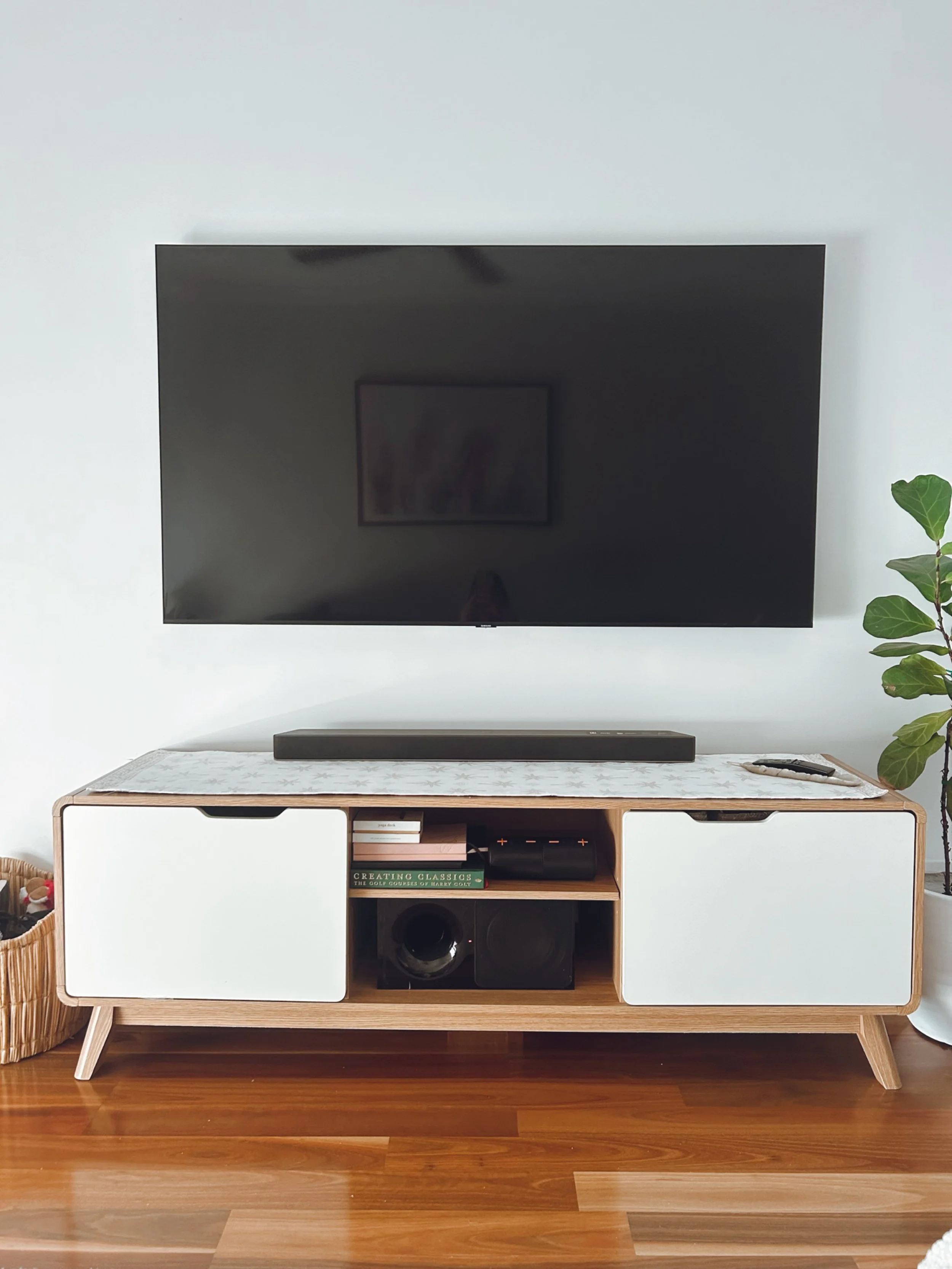 Modern entertainment center with a mounted flat-screen TV, a soundbar, and a few books, with a plant on the right and a wicker basket on the left.