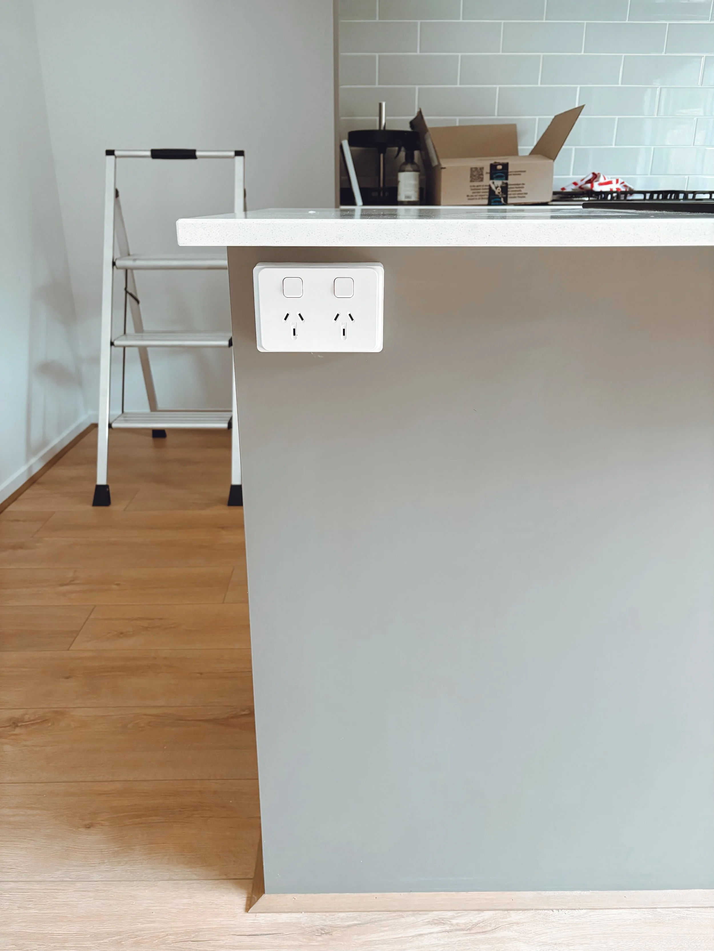 Close-up of a kitchen island with a white electrical outlet on the side, light gray base, and white countertop. In the background, there is a step ladder, a cardboard box, and some kitchen items on a counter.