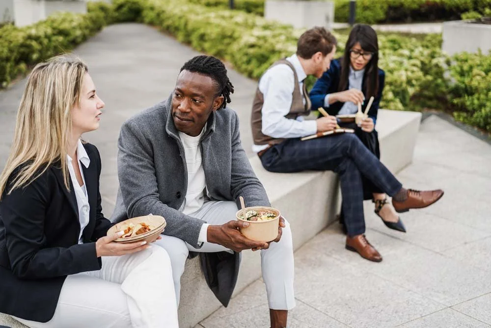 Corporate employees eating lunch on outdoor bench at landscaped headquarters, showcasing corporate landscape design that improves employee morale and workplace satisfaction.