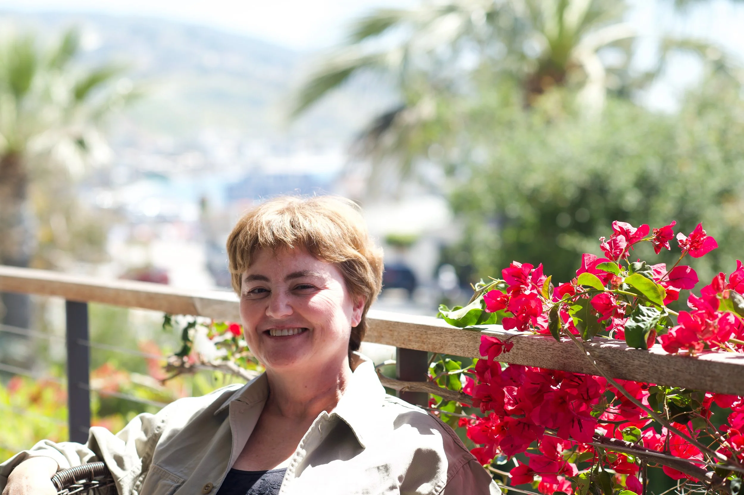 A smiling woman with short light brown hair is sitting outdoors on a sunny day, next to a bright pink flowering bush, with a blurred background of trees and water.