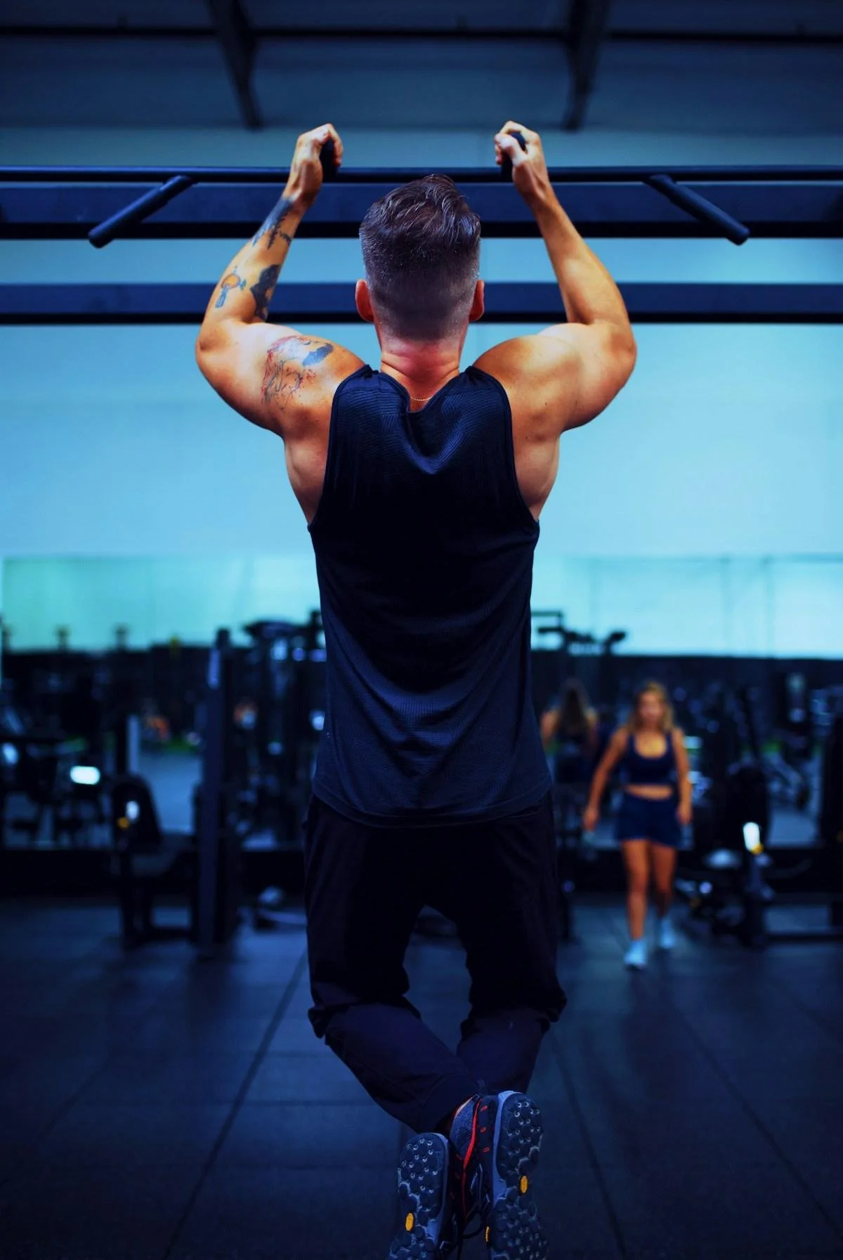 A man with tattoos wearing a black athletic tank top and pants is doing pull-ups in a gym, viewed from behind, with a woman in athletic wear walking in the background.