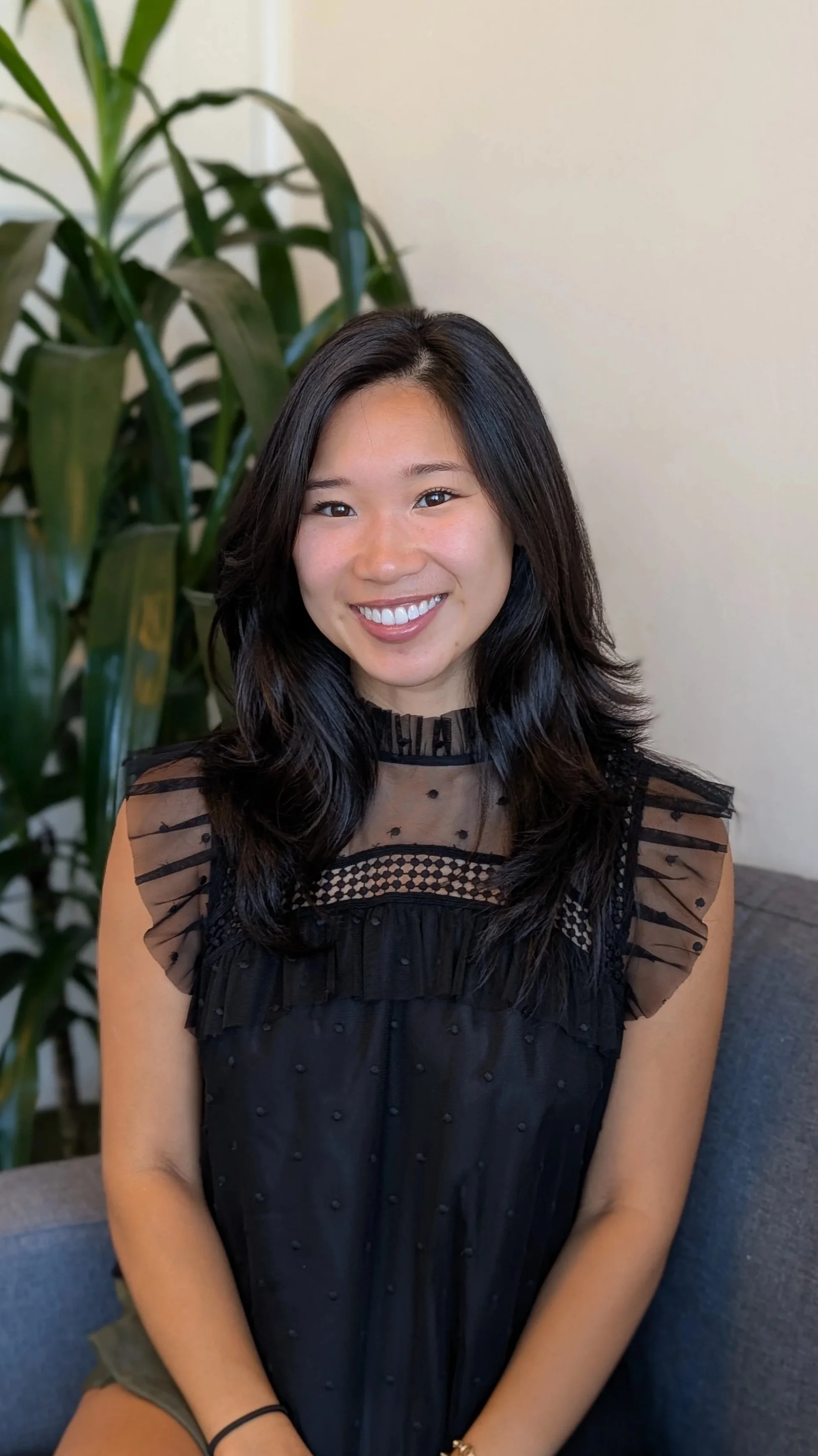 A young woman with dark hair and a bright smile seated on a blue couch, with a large green plant in the background, wearing a black lace dress.