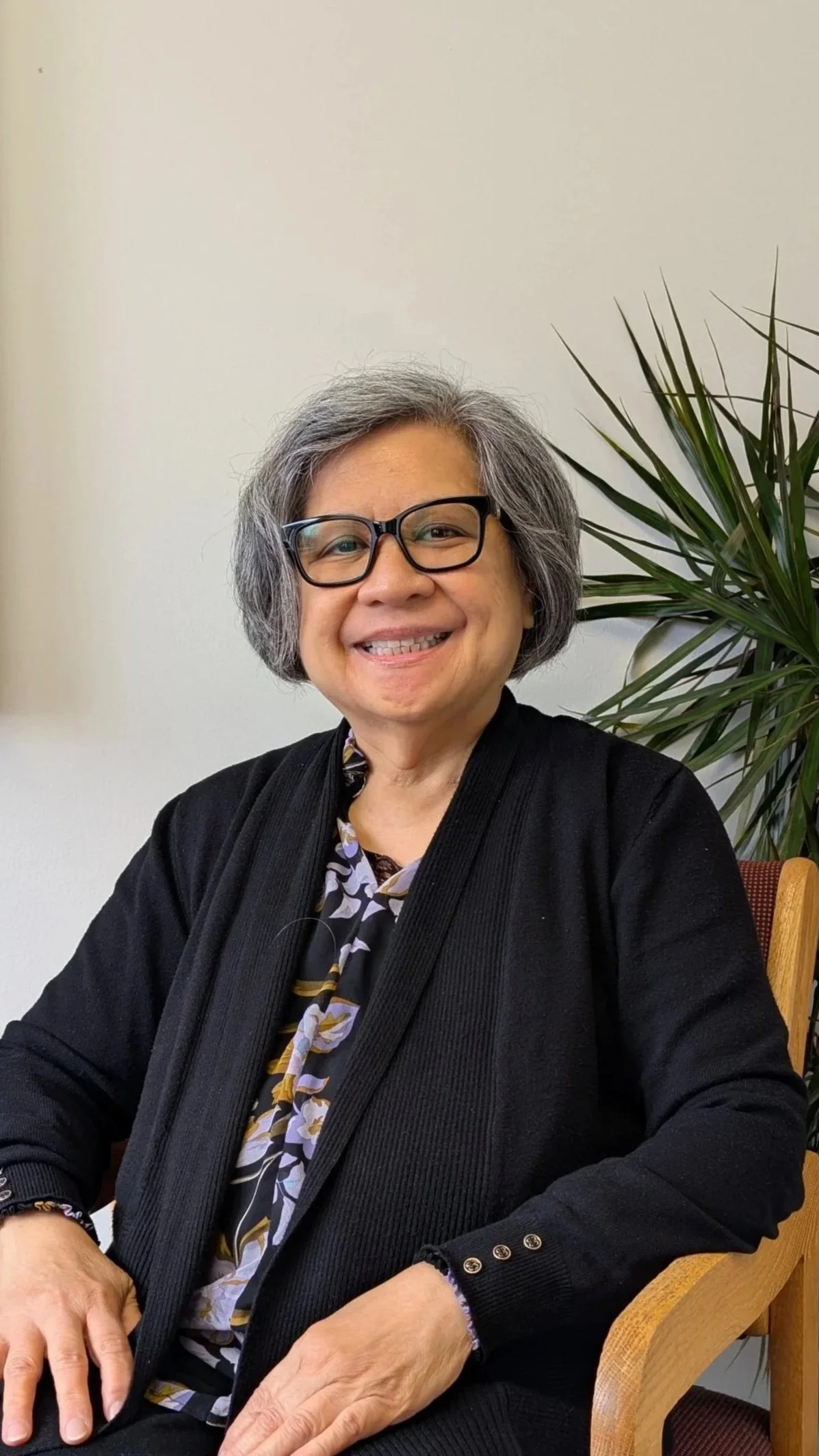 A smiling older woman with gray hair and glasses sitting next to a green plant in an indoor setting.