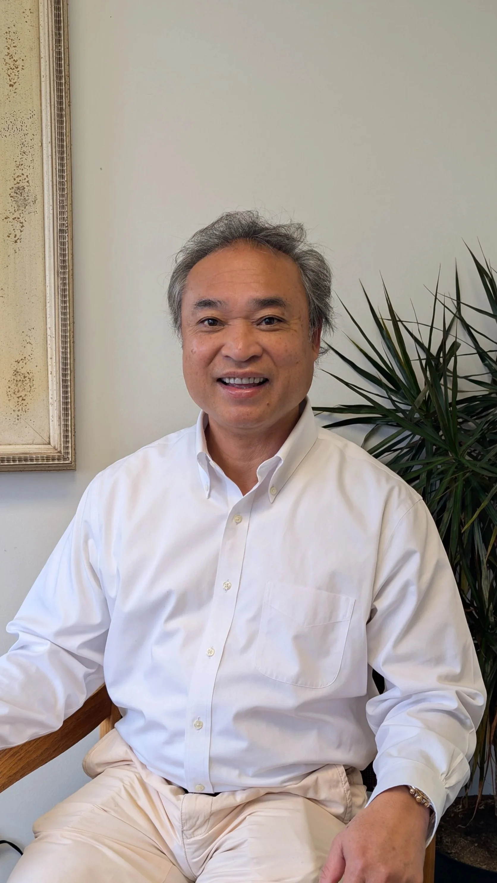 A smiling man with gray hair, wearing a white dress shirt and cream-colored pants, sitting on a wooden chair next to a plant. There is a framed piece of art on the wall behind him.