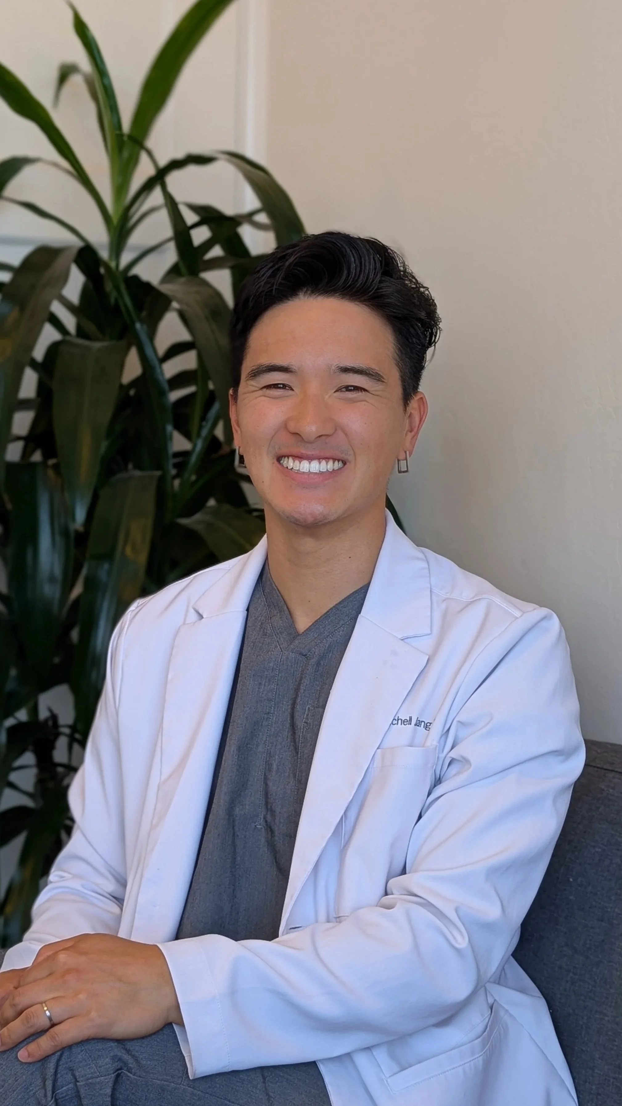 A smiling man wearing a white medical coat sitting on a gray couch with a green leafy plant in the background.