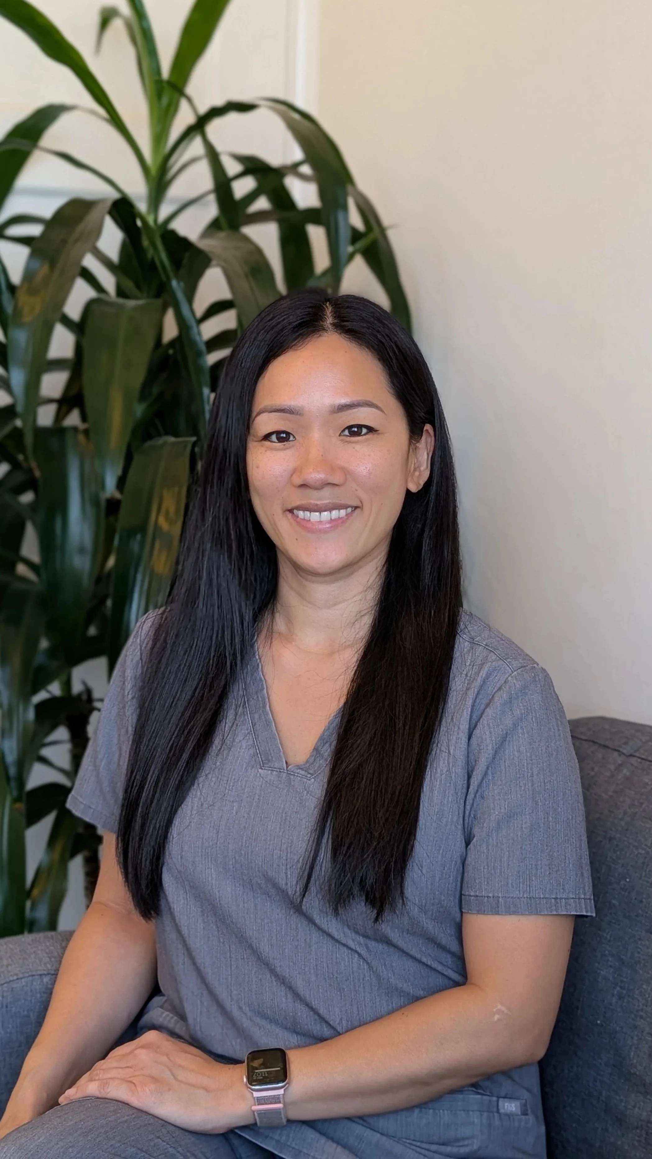 A woman with long black hair, wearing a gray scrub top, sitting on a dark gray couch with a large green plant behind her, smiling at the camera.