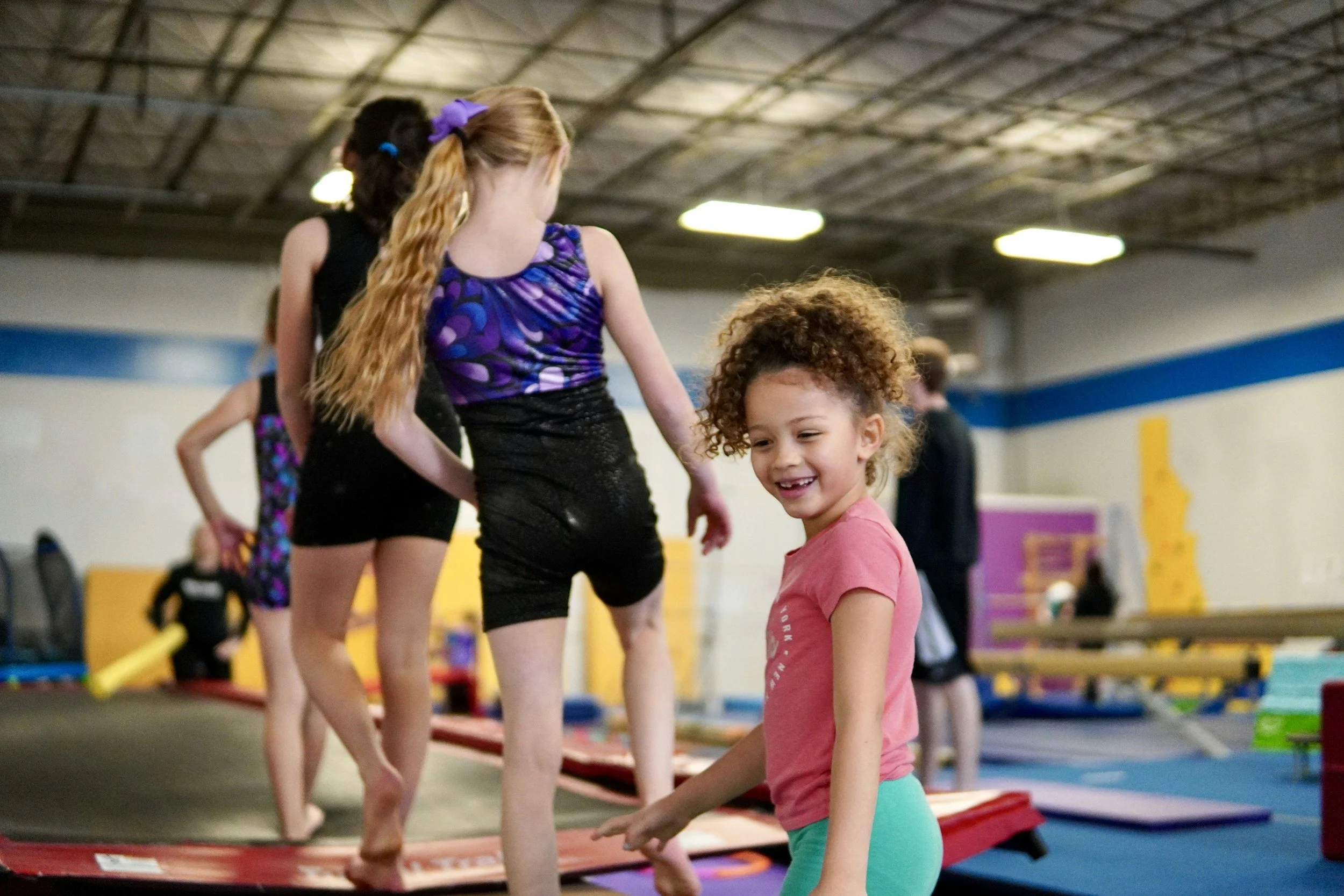 Children participating in a gymnastics class, balancing on a beam, in an indoor gymnasium.
