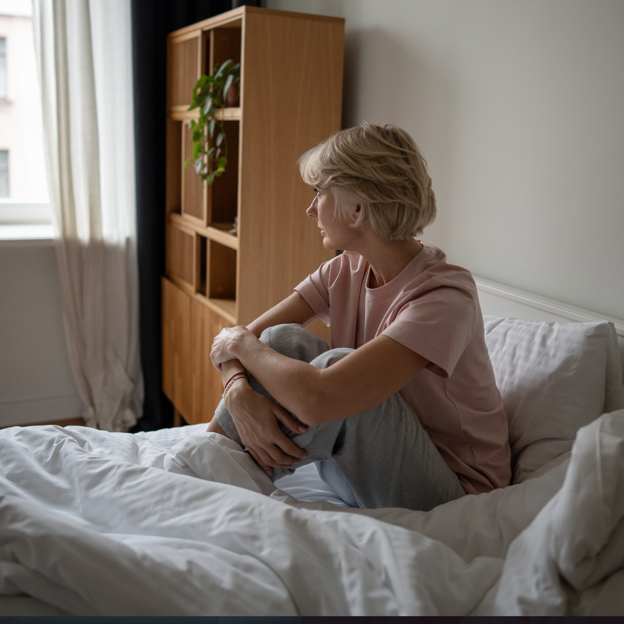 A woman sitting quietly on a bed looking out the window, representing the emotional and physical impact of fatigue and the importance of rest, pacing and occupational therapy support