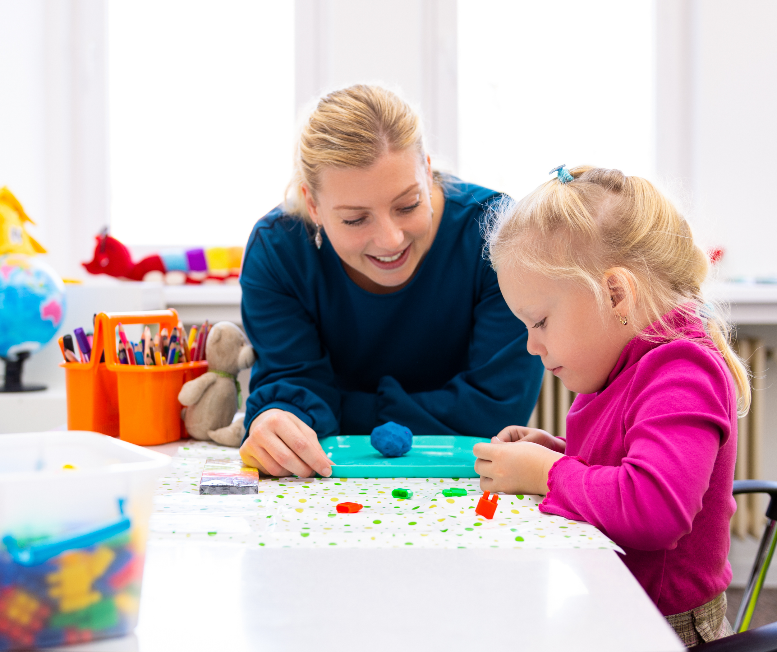 Paediatric therapist supporting a young child during a creative play activity at a table, using colourful craft materials in a bright and welcoming clinic setting.