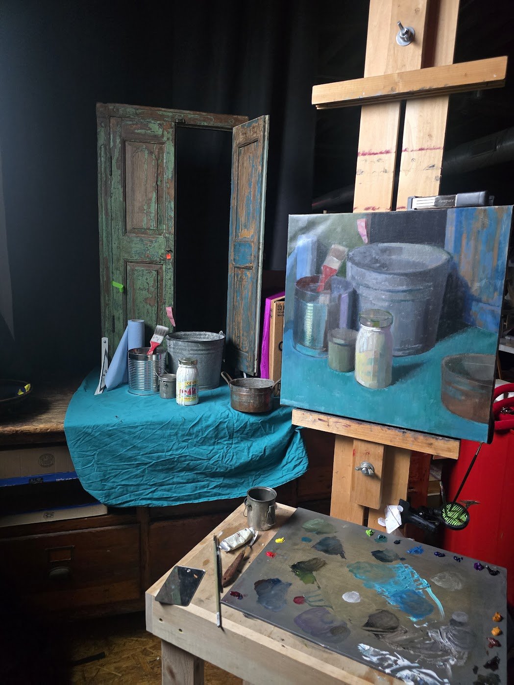 An artist's workspace with a still life painting of paint jars and containers on a canvas, a palette with mixed colors, brushes, and art supplies, and a vintage green wooden shutter in the background.