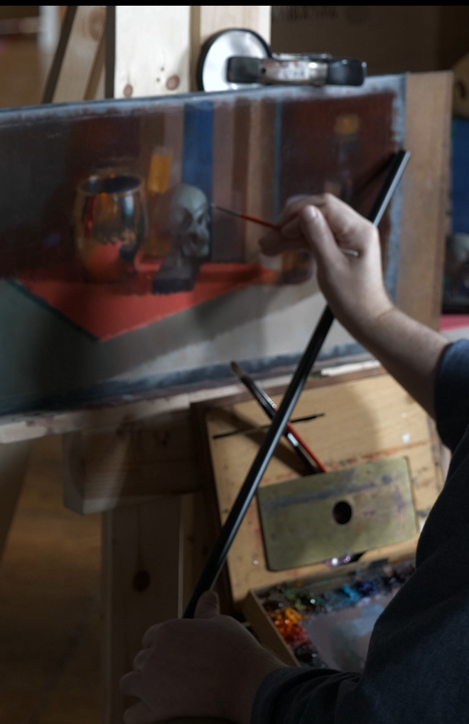 Person painting a still life with a skull, a vase, and a reflective object on a canvas in an art studio.