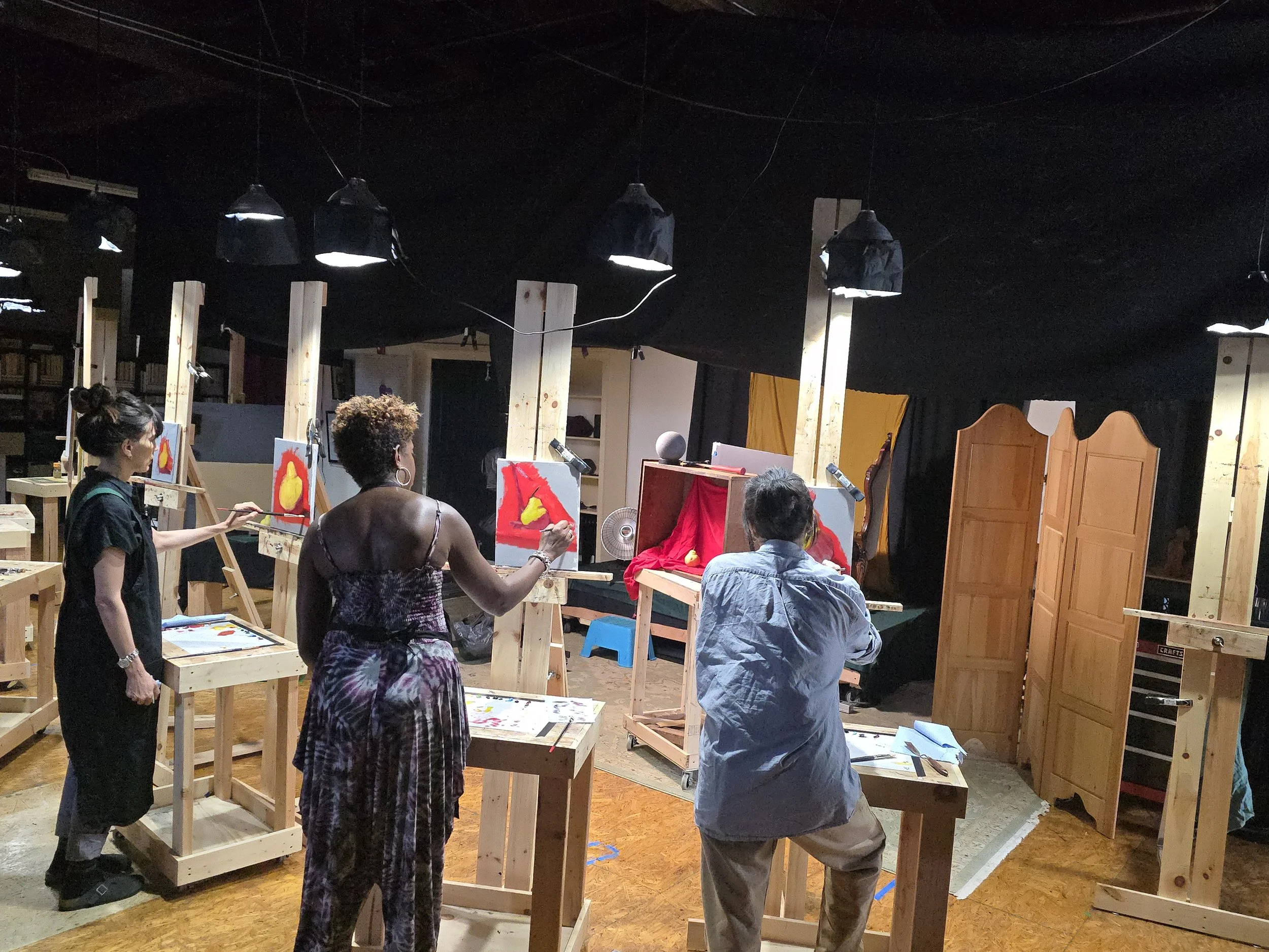 People participating in an art class, painting abstract art on canvases set on wooden easels inside a studio with black ceiling and wooden partitions.