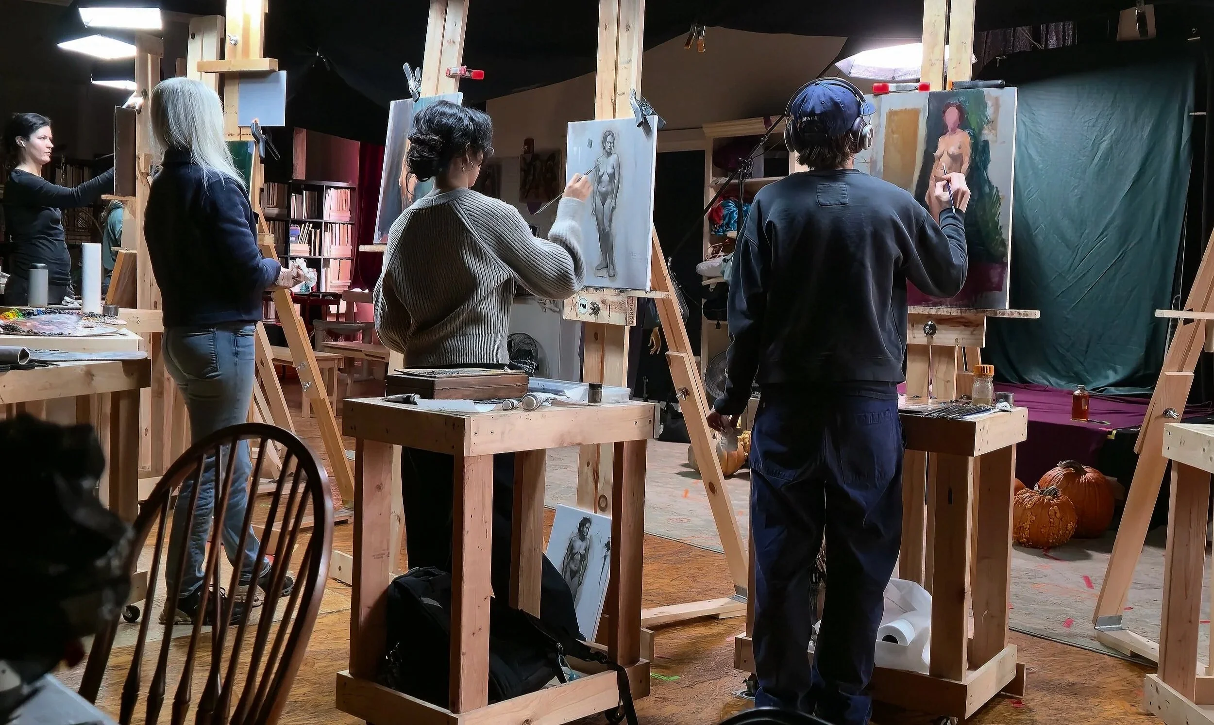 People painting in a rustic art studio with wooden easels, some finished artworks on display, and pumpkins on the floor.