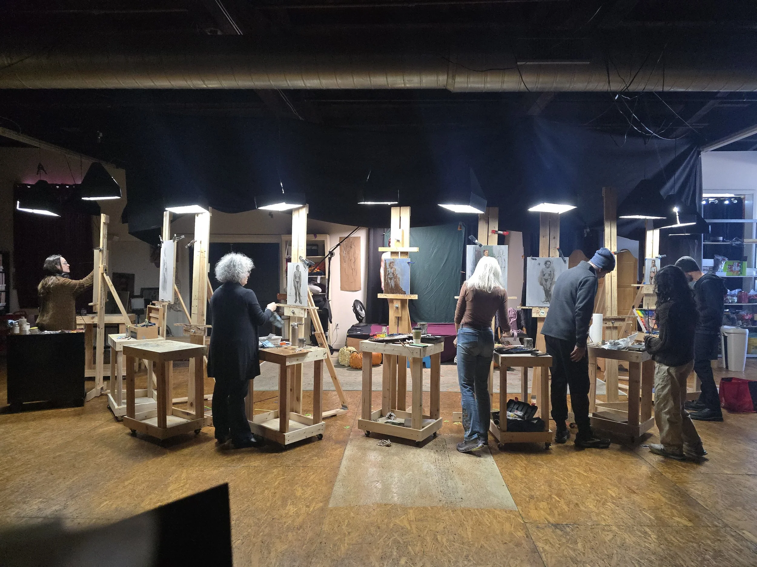 People painting on canvases set up on easels in an art studio, with small tables and studio lights overhead.