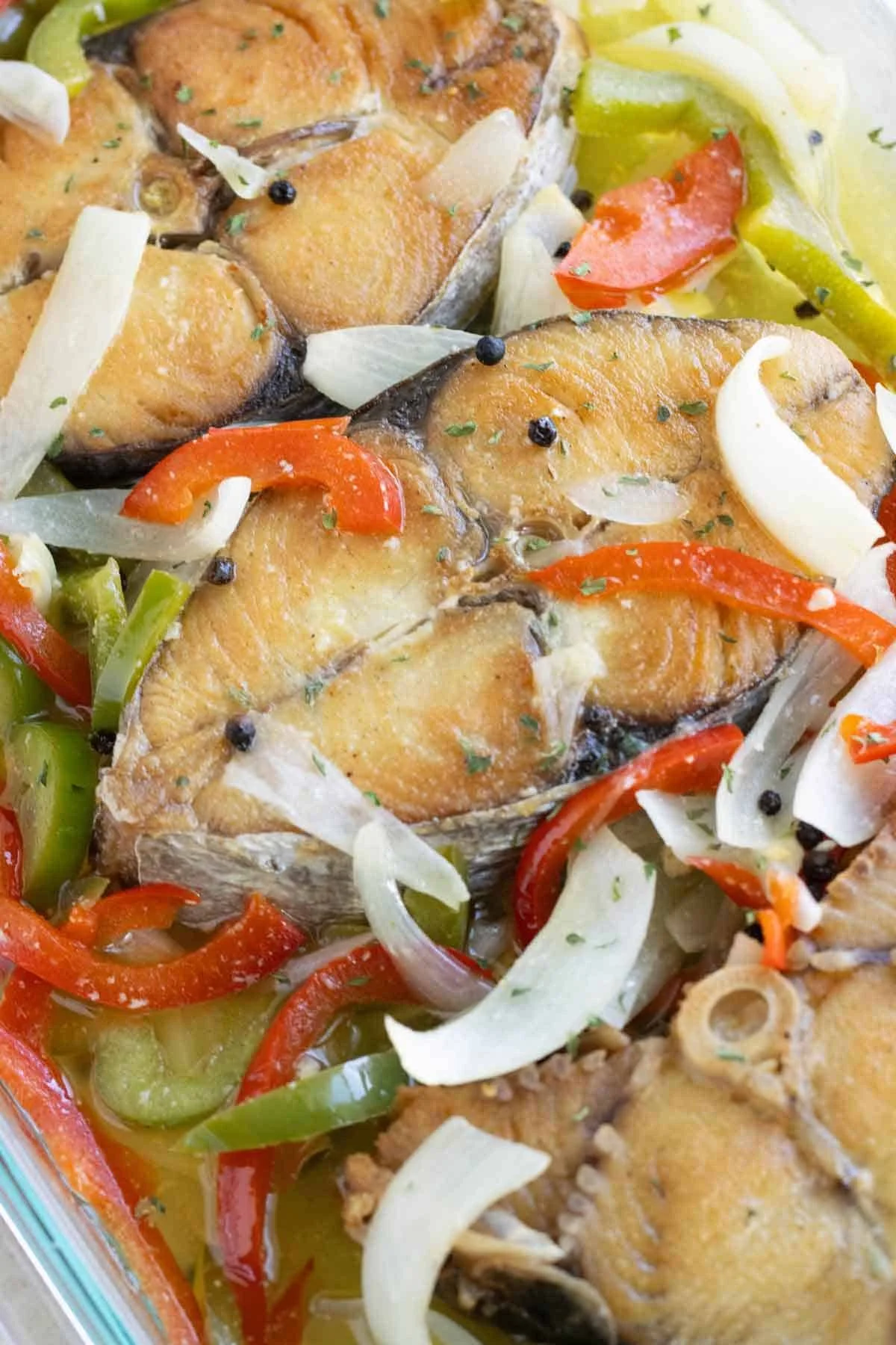 Baked fish fillets topped with sliced onions, red and green bell peppers, with black peppercorns and herbs, served with a side of cooked vegetables.