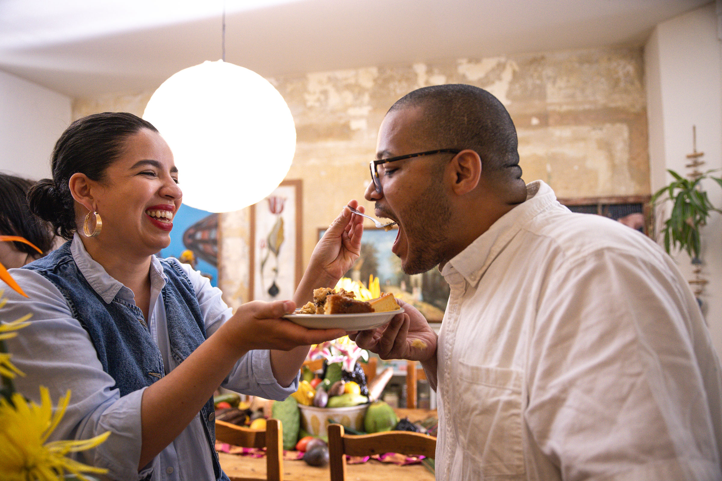 A woman is feeding a man a piece of cake at a gathering in a home with a decorated table and artwork on the walls.
