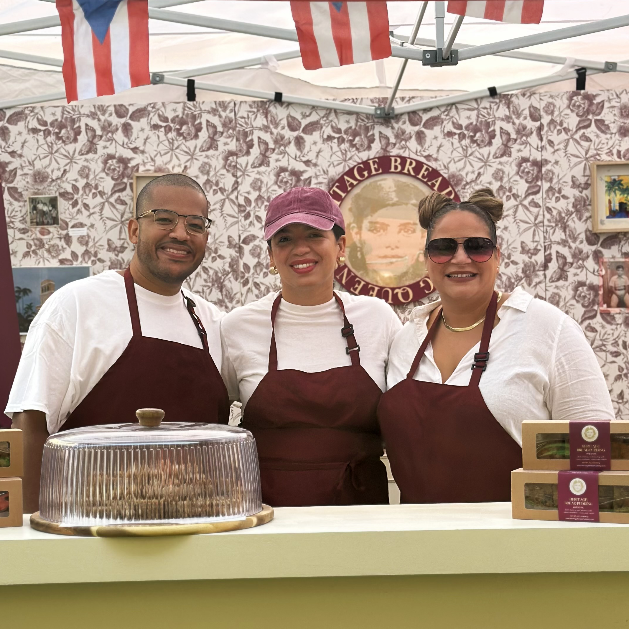 Three people wearing white shirts and maroon aprons smiling at a food booth with a banner that reads 'VINTAGE BREAD' in the background. The booth is decorated with floral wallpaper, and there are baked goods on display.