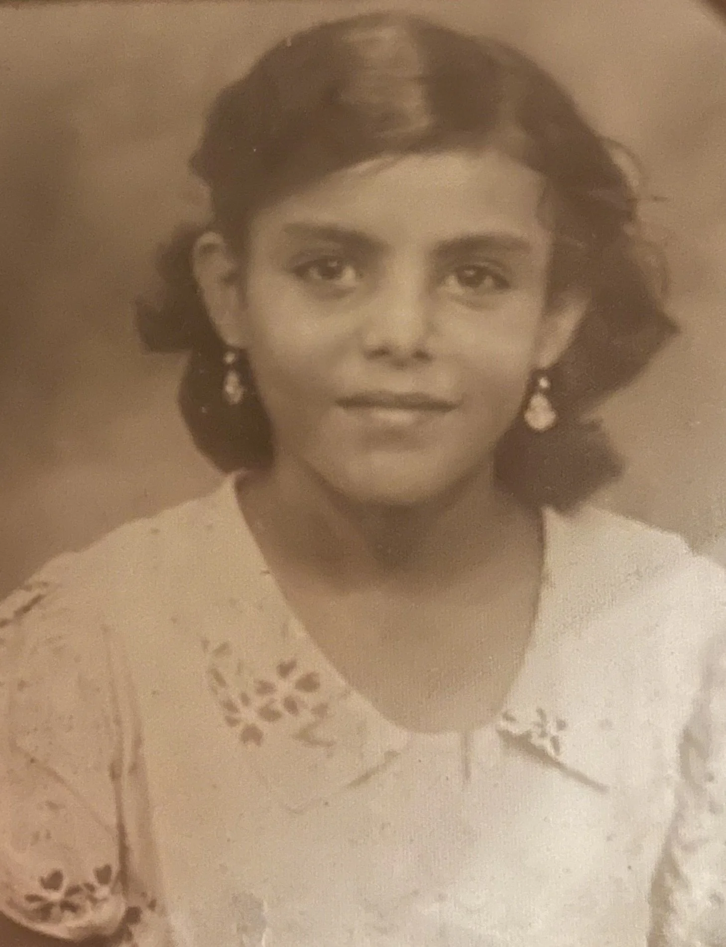 A vintage black and white portrait photograph of a woman with short, wavy hair, wearing earrings and a decorated blouse.