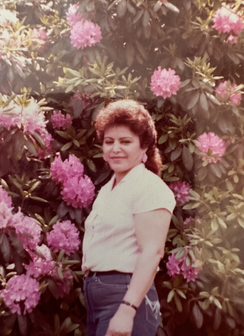 A woman with curly brown hair, wearing a white shirt and dark jeans, standing in front of pink rhododendron flowers and green foliage.