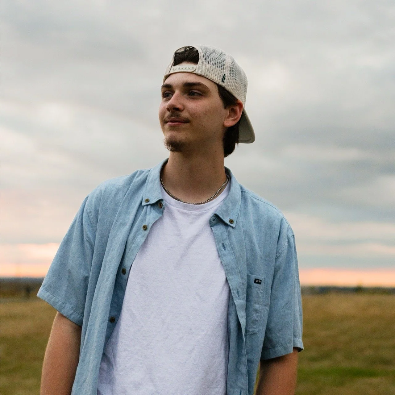 A young man with dark hair, light beard, and mustache standing outdoors in a field under cloudy sky, wearing a white baseball cap backward, white T-shirt, and a light blue button-up shirt.