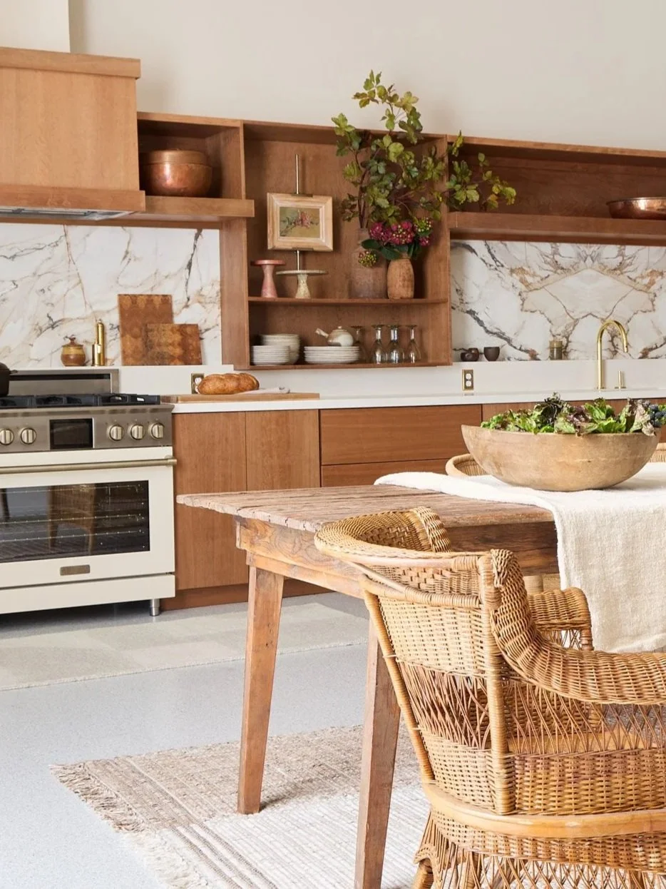 A cozy kitchen with wooden cabinets, a marble backsplash, a stove, a wooden dining table with rattan chairs, a large wooden bowl of salad, and decorative shelves with plants and dishes.