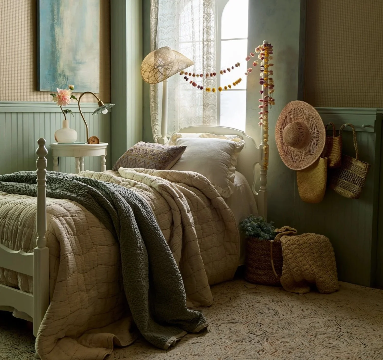 A cozy bedroom with a white bed, layered blankets, and pillows. Natural light filters through a window, decorated with hanging garlands. A bedside table with a vase and flower, and woven hats and bags hang on the wall next to a basket with blankets.