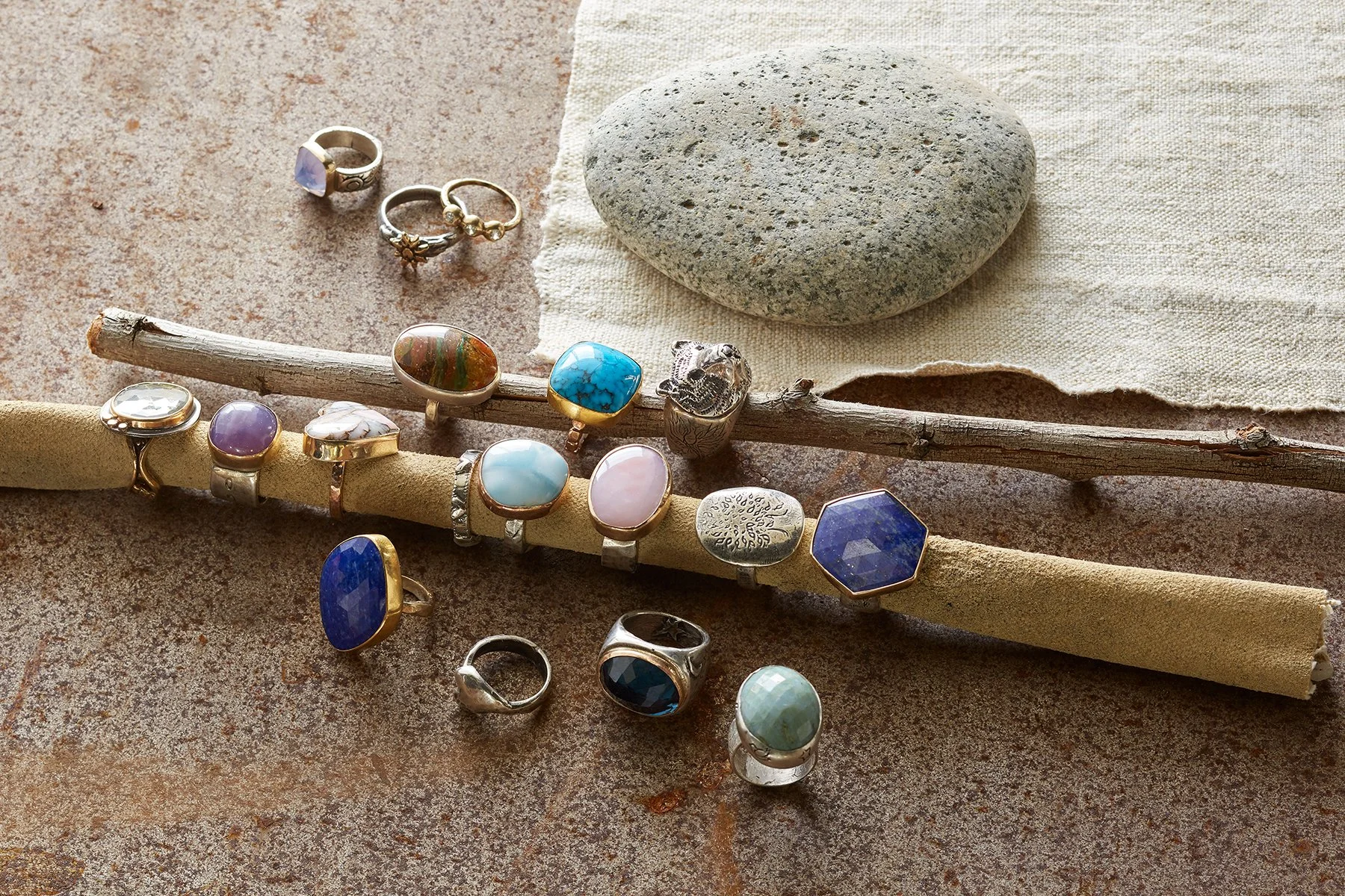 Collection of jewelry rings with various stones, a gray and a multicolored stone, on a rustic surface with rocks and twigs for decoration.