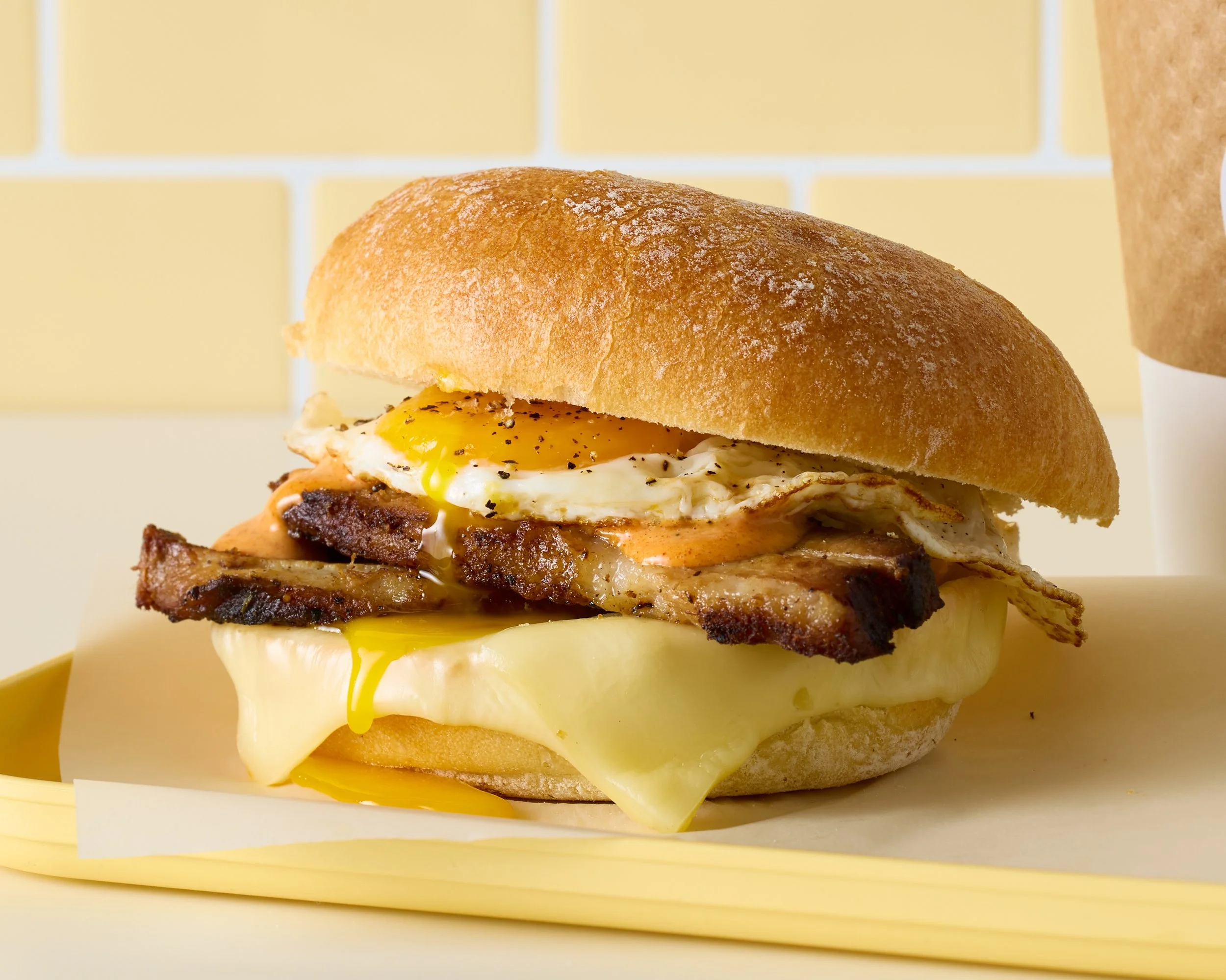 A breakfast sandwich with a fried egg, bacon, cheese, and a bun on a light yellow tray.