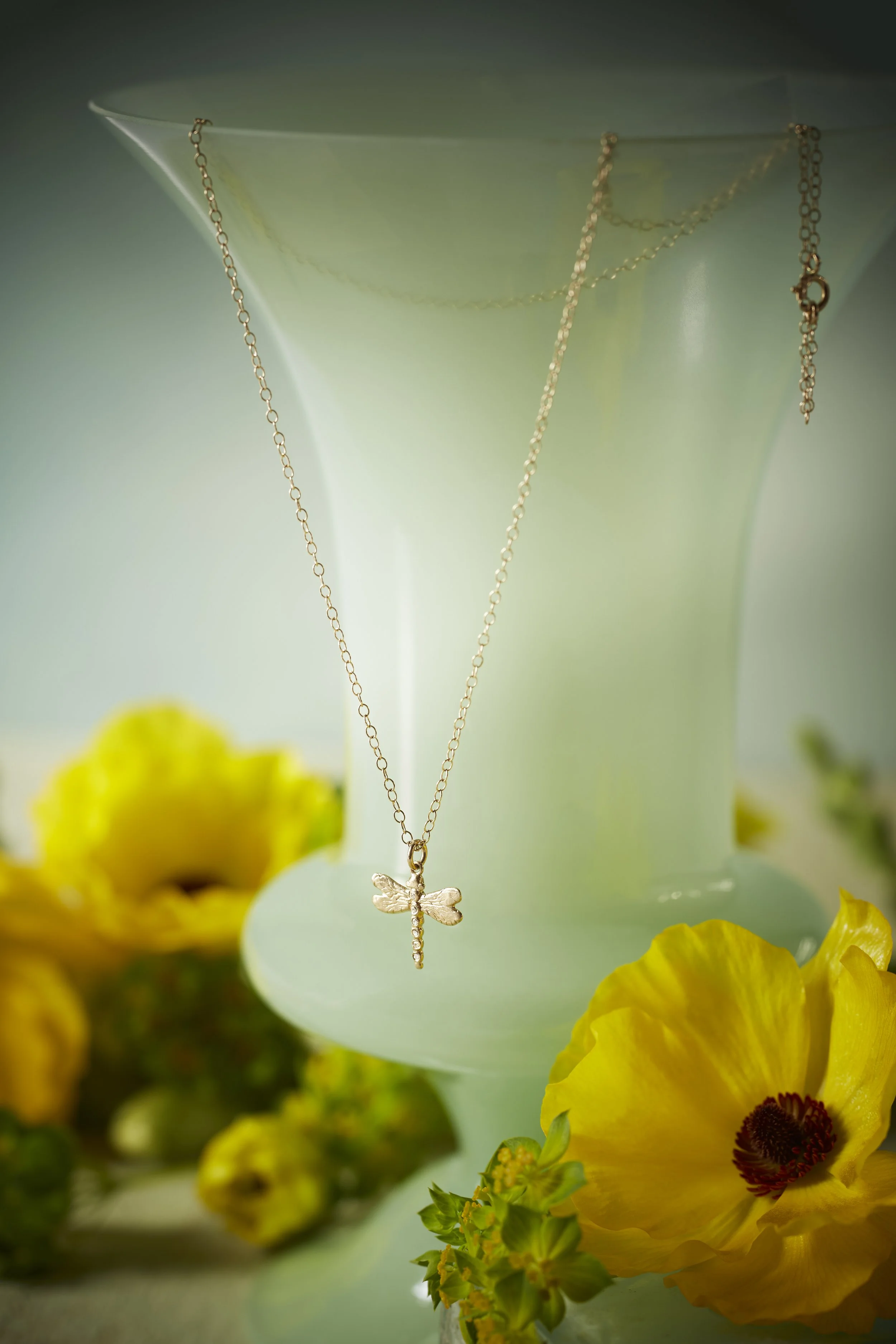 A gold dragonfly pendant necklace hanging from a light-green vase with yellow flowers in the foreground.