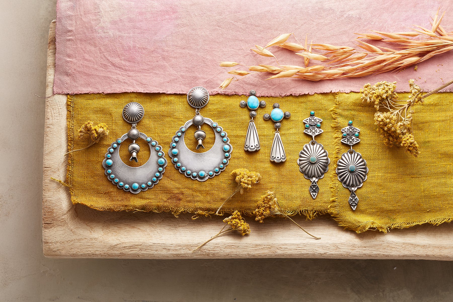 Six pairs of silver earrings with turquoise accents displayed on yellow fabric, with dried flowers and pink fabric in the background.