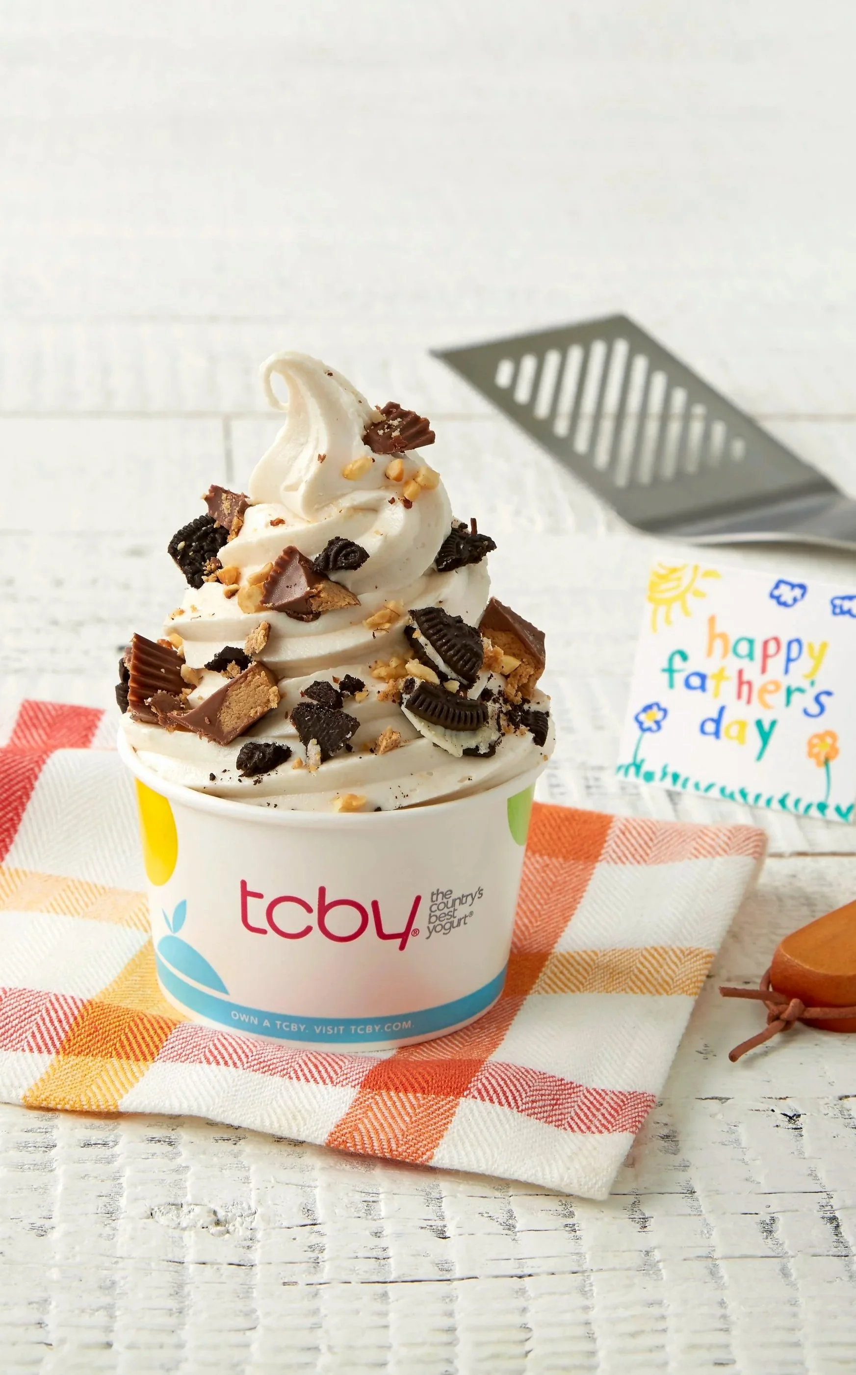 A cup of vanilla frozen yogurt topped with chocolate and cookie crumbles, on a checkered cloth with a Father's Day card in the background.