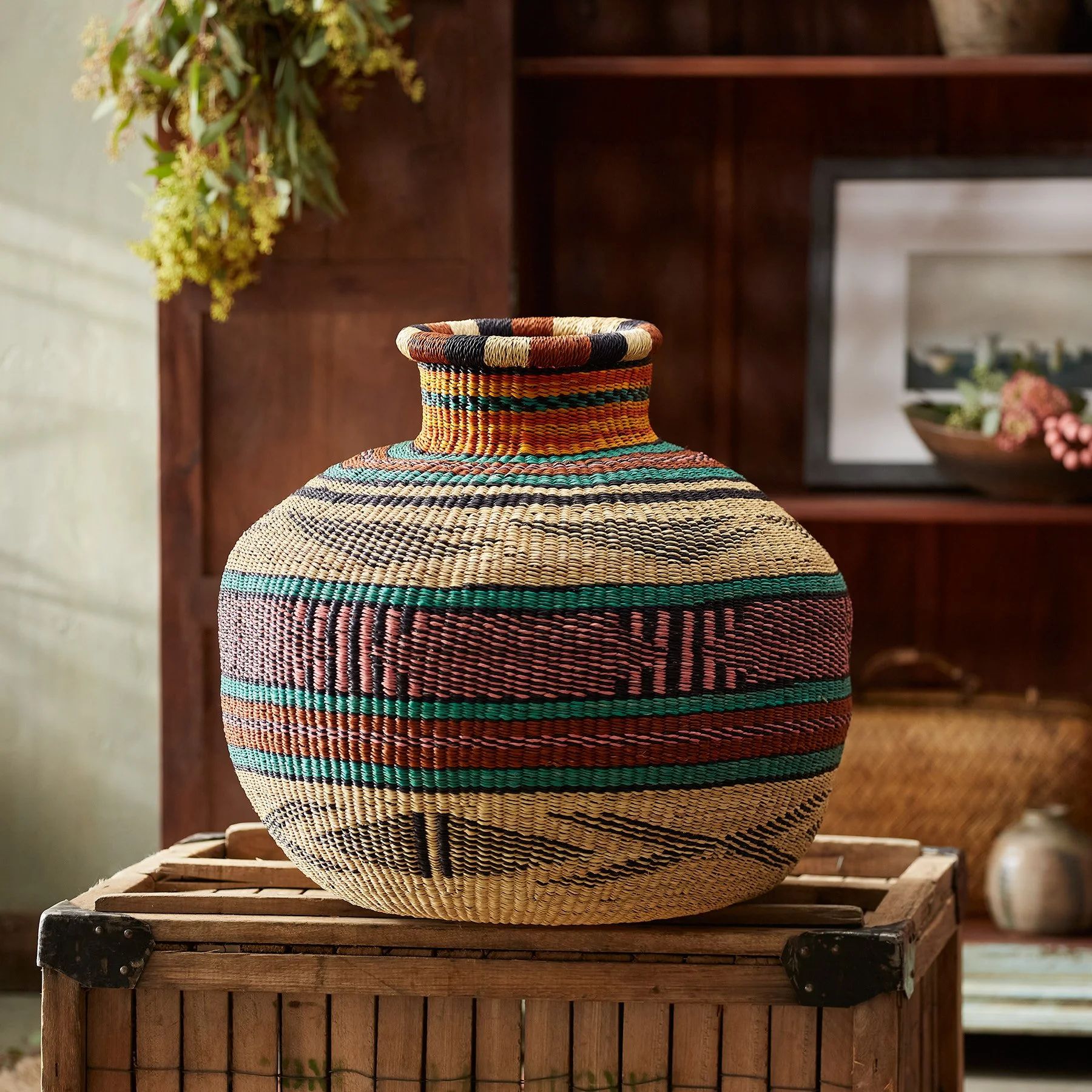 Colorful woven basket with geometric patterns on a wooden stand in a room with wooden shelves and framed photos.