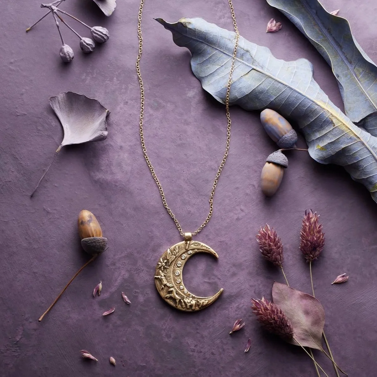 A gold necklace with a crescent moon pendant resting on a purple surface, surrounded by dried foliage, acorns, and leaves.