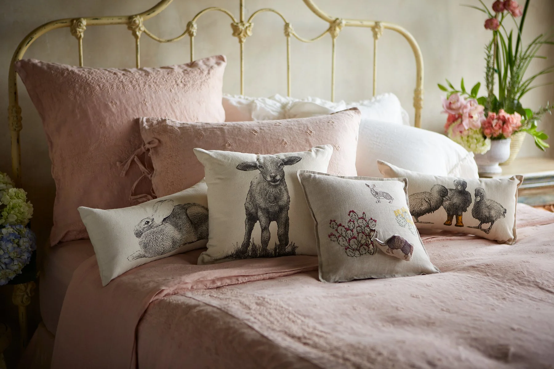 A bed with a light pink quilt, multiple pink pillows, and decorative pillows with animal and botanical embroidery, a floral arrangement in a white vase on a side table, and greenery in the background.
