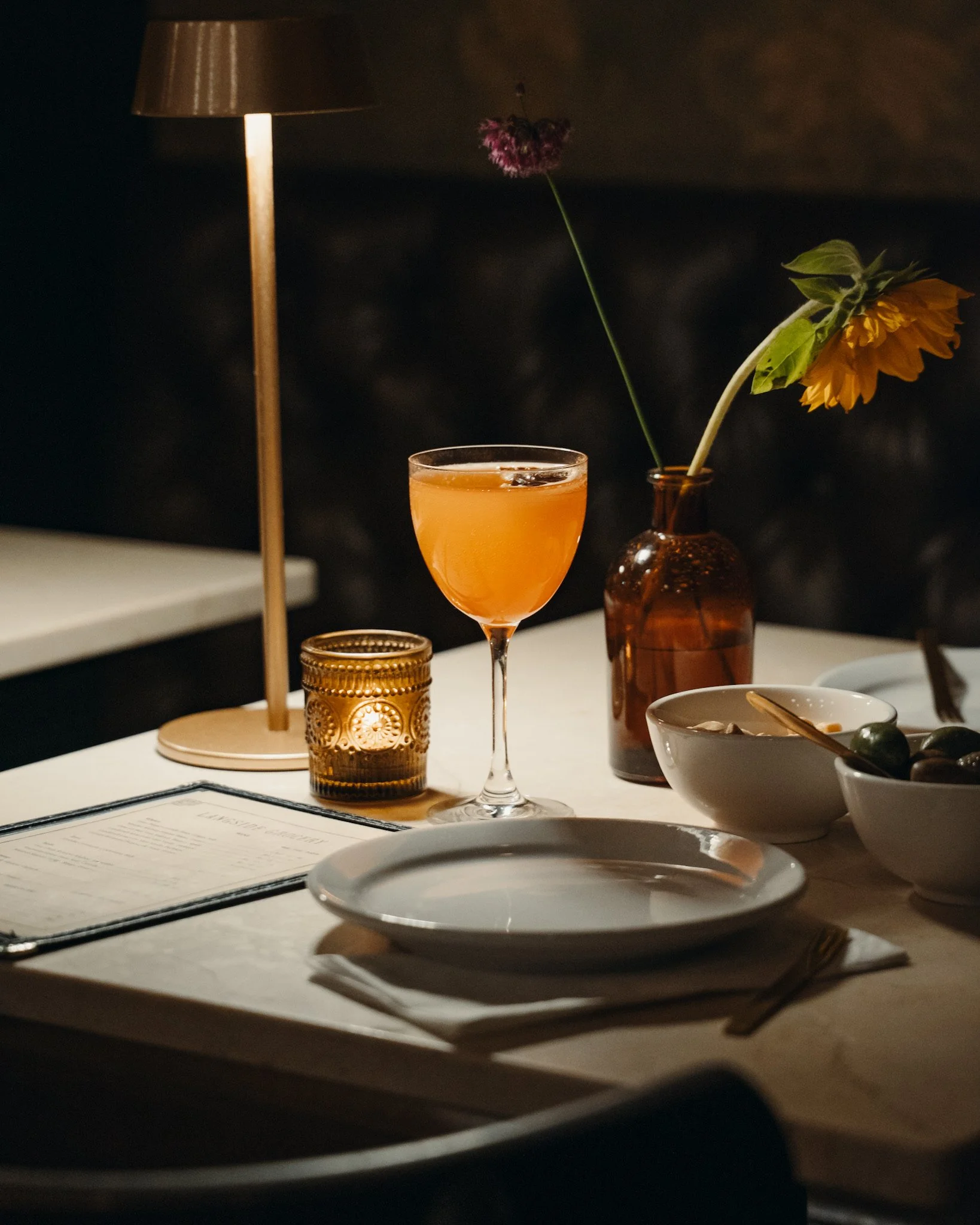 A dimly lit dining table features a cocktail glass with an orange-colored drink, a brown glass vase with a purple flower and a yellow flower, a small amber candle holder, a white bowl with a spoon, and a menu.
