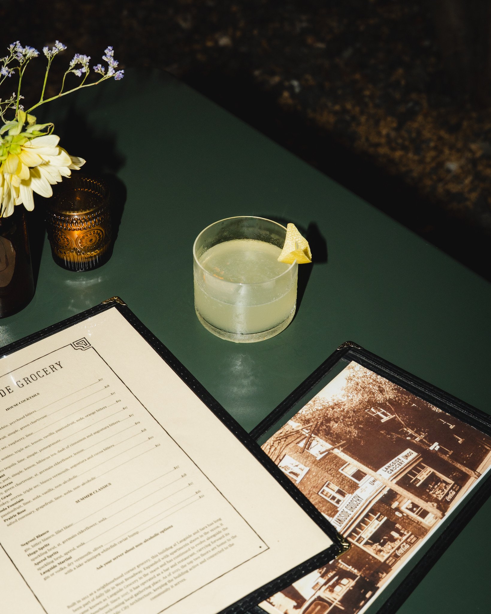A menu on a table with a floral arrangement, a candle holder, and a cocktail with a lemon wedge. The menu features sections for grocery, house cocktails, summer classics, and a vintage photo of a building.