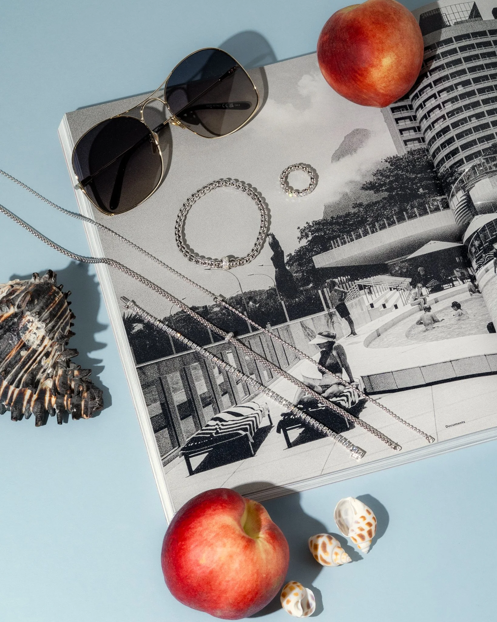 A flat lay of two red apples, a pair of sunglasses, a silver bracelet, a silver necklace, four small seashells, and a seashell with a dark, textured exterior, all placed on an open black-and-white photo of an urban park with trees and buildings.