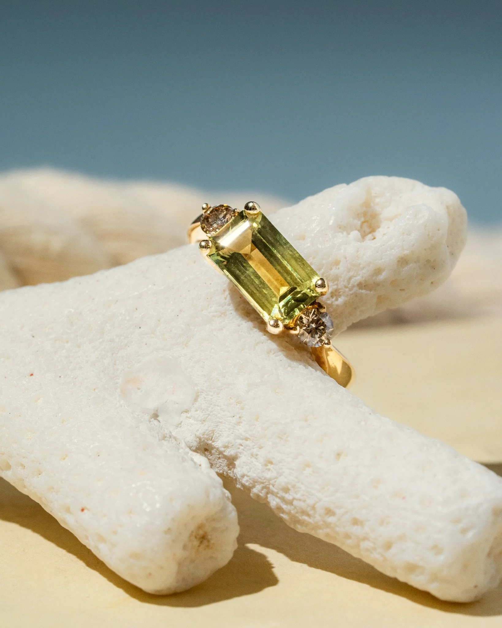 A gold ring with a rectangular yellow gemstone and two small clear stones, resting on white coral-like rocks.