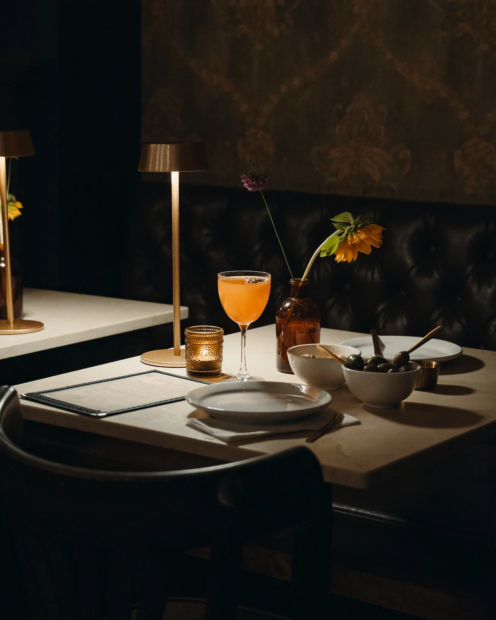 A cozy dining table setup in a dimly lit restaurant, featuring a glass of orange-colored beverage, a small bowl with olives, a bowl of sauce, a fork, a knife, and a menu, with two table lamps providing warm lighting.