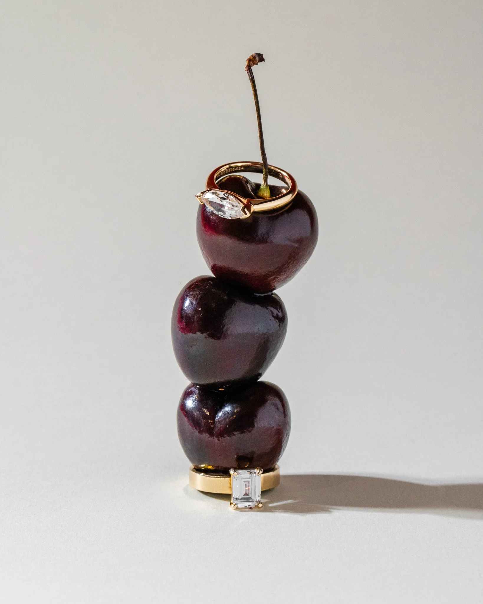 A stack of four dark cherries with glossy skin, topped with a gold ring featuring a large, marquise-cut clear gemstone, and a single emerald-green leaf stem on the cherry at the top. The ring is placed around the cherry's stem, with the gemstone resting on the cherry's surface.