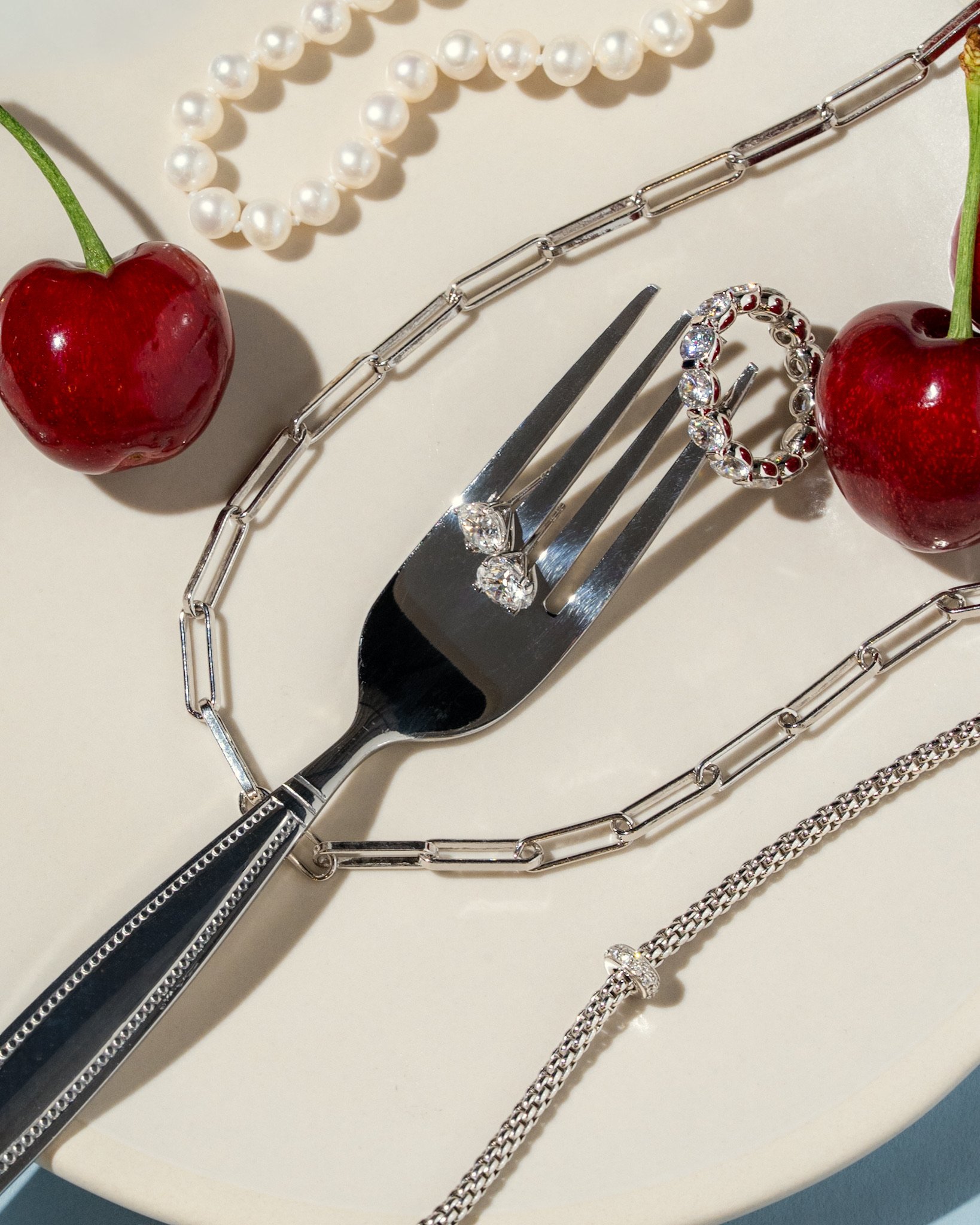 Jewelry and accessories on a white surface, including a pearl necklace, two cherries, a silver chain, a pair of high-heeled shoes with rhinestones, and a diamond ring.