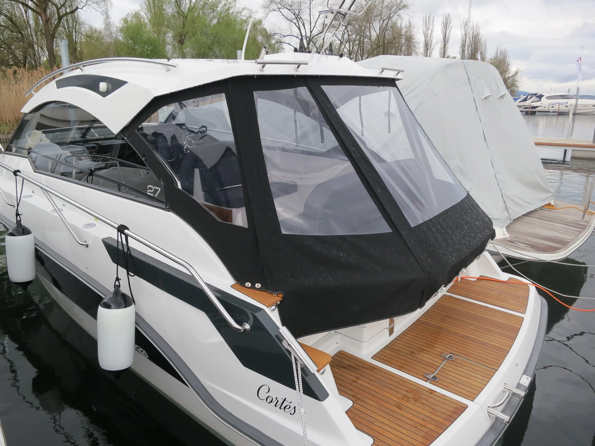 Ein modernes Motorboot im Hafen mit abdeckbarer Kabine, Fender an den Seiten und Holzplattform am Heck. Es liegt neben einem weiteren verhüllten Boot.