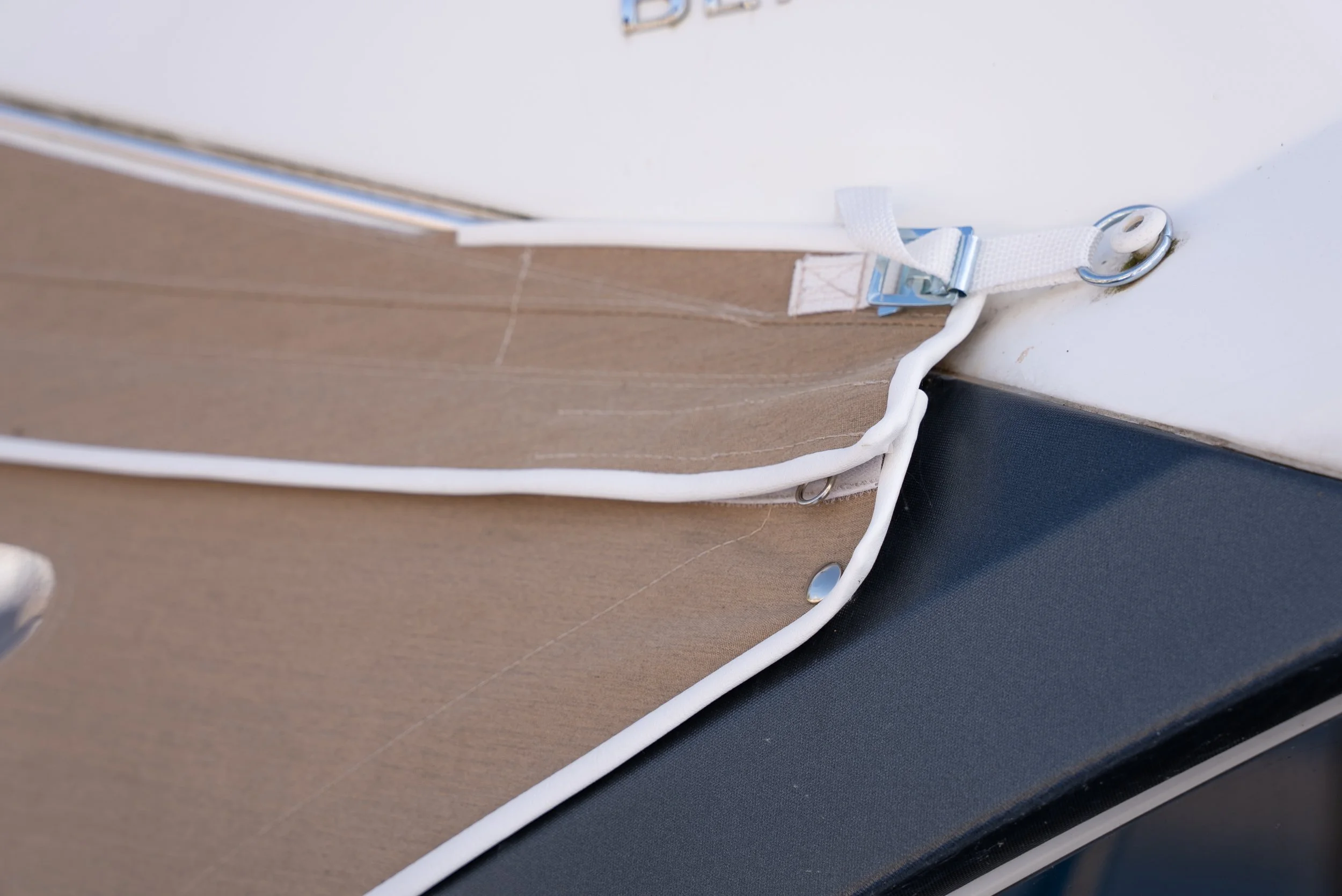 Nahaufnahme einer Plane aus Stoff mit einem Befestigungsgurt und Metallösen, die an einer harten Oberfläche befestigt sind. Die Kanten der Plane sind mit einem weißen Band verstärkt.