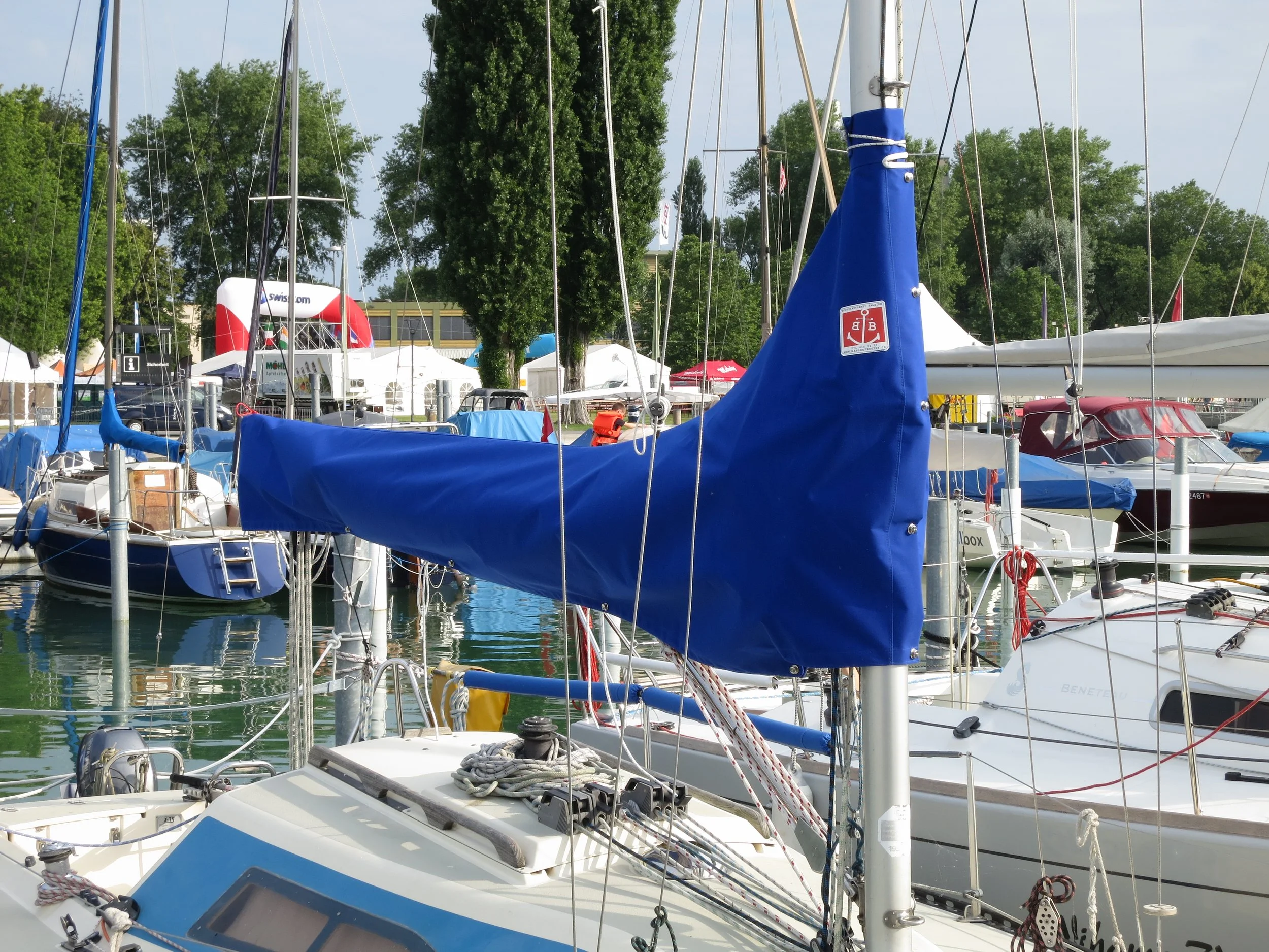 Yachten im Hafen mit blauen Segelabdeckungen, umgeben von Bäumen und Zelten.