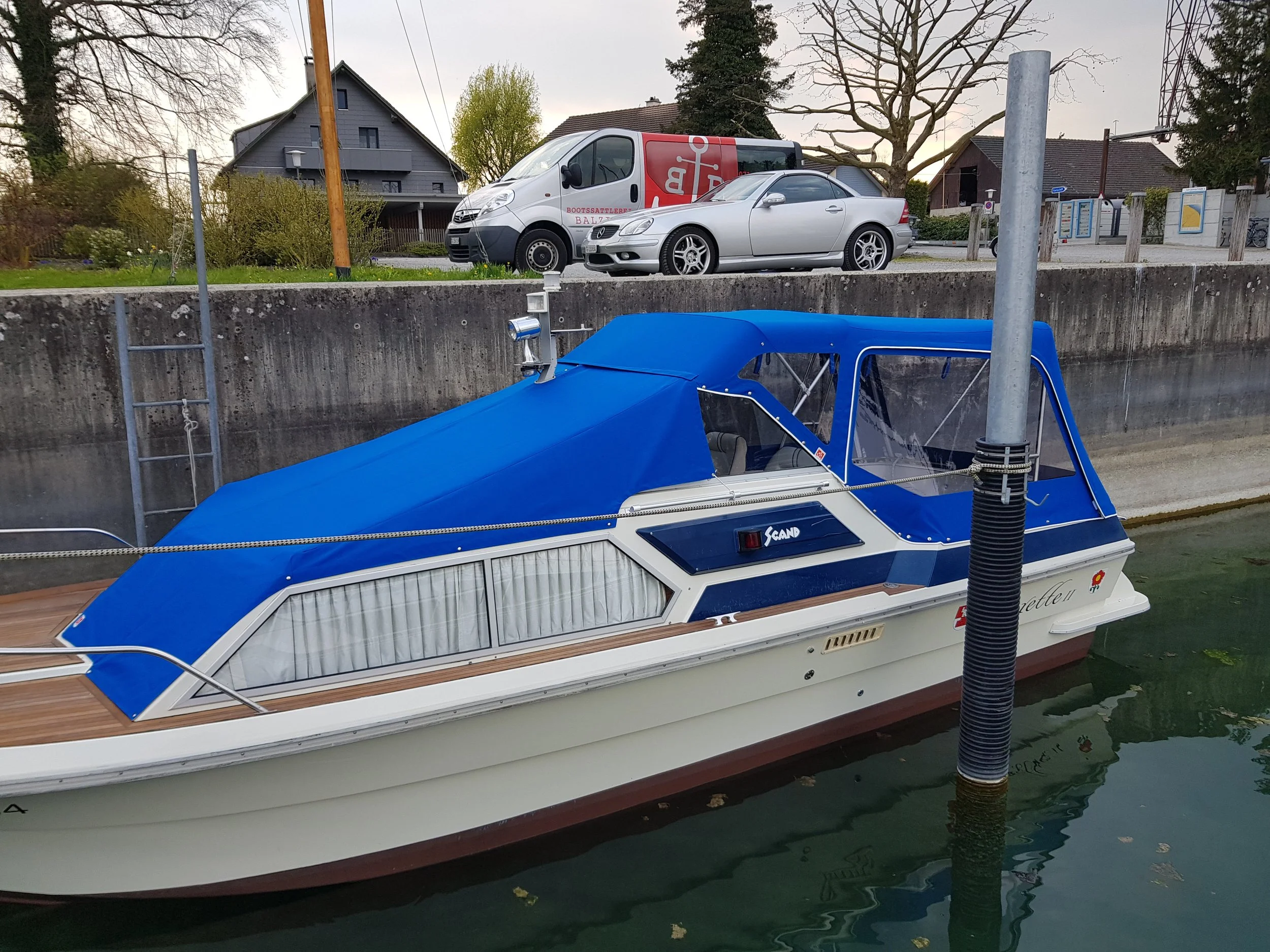 Ein Motorboot mit blauer Abdeckung liegt im Wasser. Im Hintergrund sind eine Betonmauer, eine Leiter, ein Lieferwagen und ein Auto zu sehen. Es gibt auch ein Haus und Bäume.