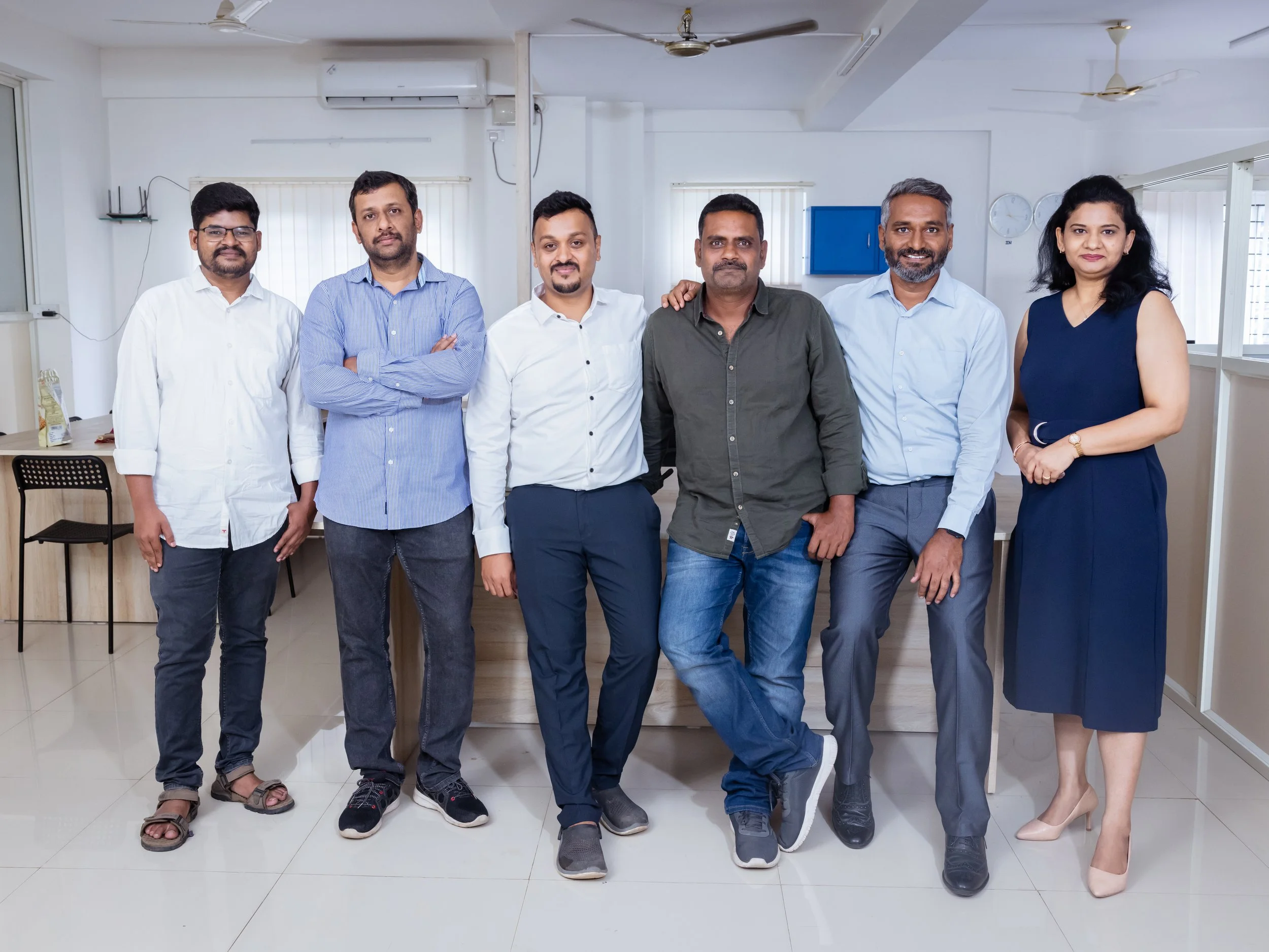 Teamfoto van zeven mensen in een kantoor, zeven mannen en één vrouw, verschillende outfits, alle lachend voor een witte muur en vensters.