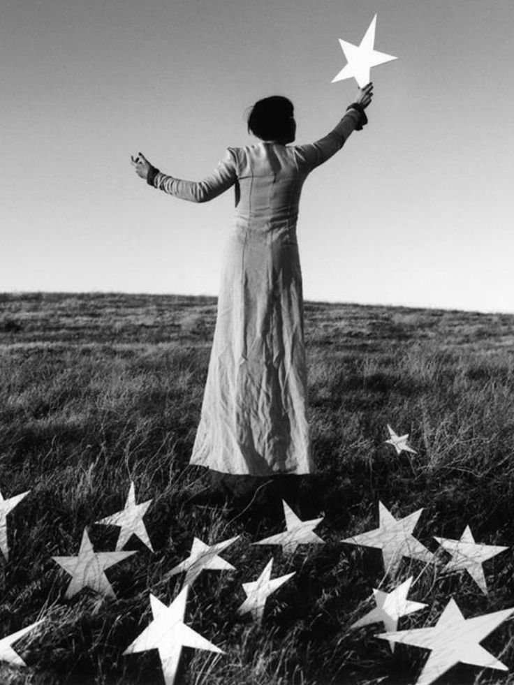 Una donna in un campo aperto che tiene in mano una stella di carta e ci sono altre stelle di carta sparse a terra, in bianco e nero  | Romina Felici - L'Officina dell'Io®